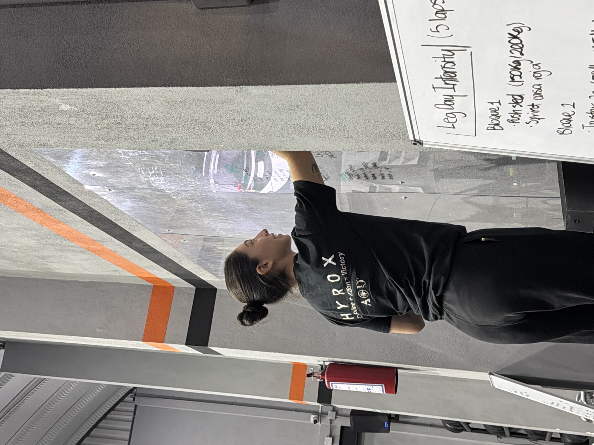 The image, appearing rotated, shows a woman in a gym setting, attentively looking upwards at a whiteboard displaying a workout routine. She wears a black short-sleeved t-shirt with the "HYROX | Discipline + Effort = Victory" logo and "AD" printed below it, paired with black athletic pants. Her dark hair is pulled back into a neat bun.
The whiteboard, affixed to a light gray wall, details a "Leg Day Intensity" workout, specifying "5 laps." Under "Bloque 1," exercises listed are "Push sled (150kg/200Kg)" and "Sprint casa roja." The heading for "Bloque 2" is also visible.
The gym's interior features robust structural beams in a light gray color. The walls are adorned with striking black and orange stripes, some diagonal and some horizontal, creating a dynamic visual. A red fire extinguisher is prominently mounted on one of the gray beams. The overall ambiance suggests a functional and well-equipped fitness facility, located in San Bernardino Tlaxcalancingo, Mexico.