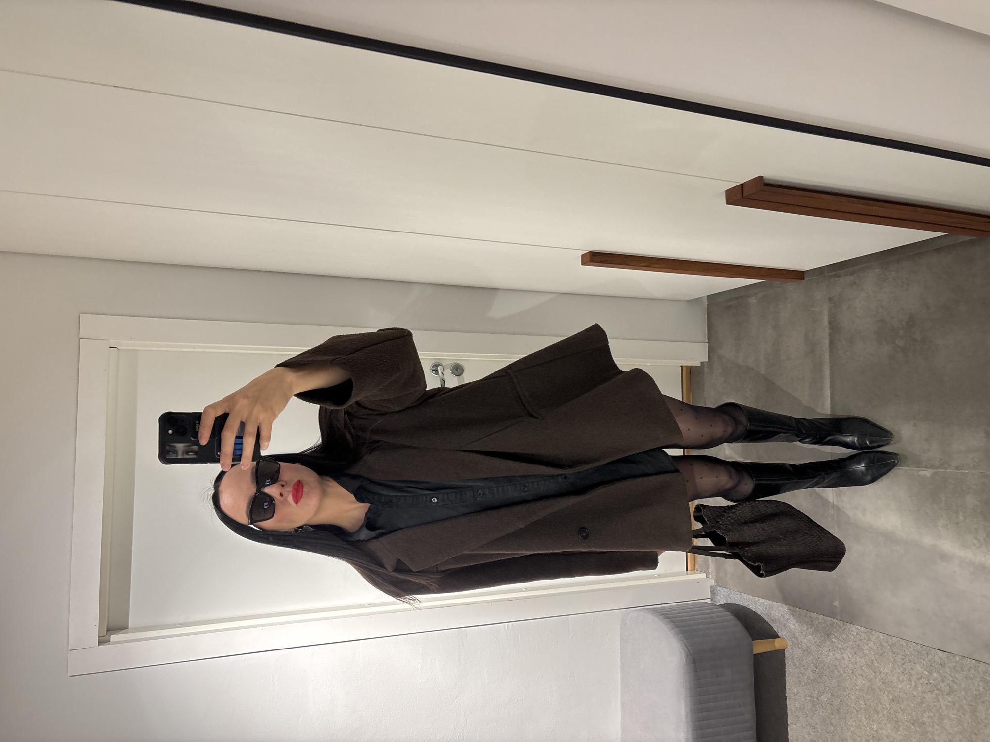 An upright mirror selfie captures a stylish woman in an indoor setting, likely in Chaikovsky, Russia. The woman, with long dark hair, is wearing a dark brown, possibly wool, coat over a dark shirt, which appears to be denim. She has sheer dark tights with a subtle polka-dot pattern and black knee-high boots. Her face is partially obscured by dark sunglasses, but striking red lipstick is visible. She holds a black smartphone with a textured case to take the photo. A dark brown, textured handbag is slung over her left arm.
The background reveals a modern, minimalist interior. The walls and ceiling are white, featuring horizontal paneling details and a prominent black trim line along the top of the ceiling. Contemporary wooden architectural elements, possibly decorative beams or shelves, are mounted on the ceiling or upper wall. The floor is a dark grey, resembling concrete. A white door frame is visible behind the woman, and a light grey upholstered, ribbed furniture piece, possibly an ottoman or bench, is in the foreground to the right. The lighting appears to be artificial and even, typical of an indoor space. No visible text is present in the image.