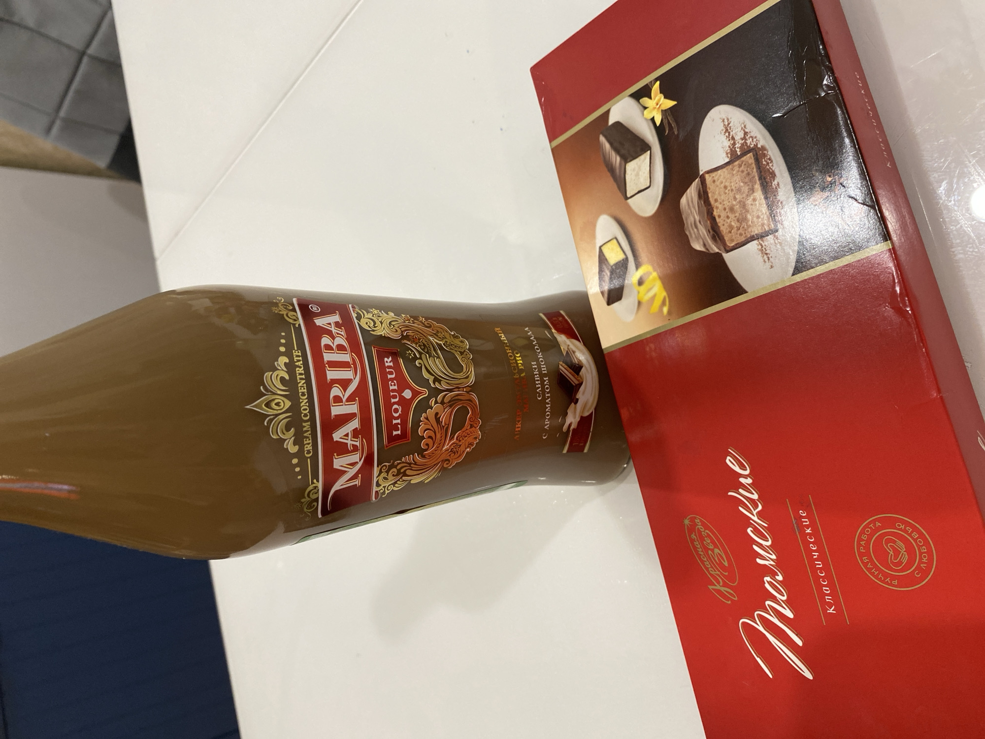 The image captures two consumer products, a bottle of liqueur and a box of chocolates, placed on a white tabletop.
The bottle, crafted from brown glass, contains a light brown, opaque liquid. Its label is predominantly red and gold, featuring the brand name "MARIBA" prominently in red script. Above "MARIBA," the text "CREAM CONCENTRATE" is visible, and below it, "LIQUEUR." Further down, Cyrillic text identifies it as a "Dessert Liqueur" (ЛИКЕР ДЕСЕРТНЫЙ) with "Cream and Chocolate Aroma" (СЛИВКИ С АРОМАТОМ ШОКОЛАДА). An illustration on the label depicts a piece of chocolate and a splash of cream.
Beside the bottle is a rectangular, red box of chocolates. The lid features ornate white Cyrillic script reading "Тюменские Классические" (Tyumenskie Klassicheskie), which translates to "Tyumen's Classic," indicating the product's origin from **Tyumen, Russia**. A smaller inscription "Красная Звезда" (Krasnaya Zvezda - Red Star) is located at the top of the lid, and a circular emblem states "РУЧНАЯ РАБОТА С ЛЮБОВЬЮ" (Handmade with Love). The right side of the box displays an image of three distinct chocolate-covered confections with different fillings (one yellow, one white, one brown), garnished with lemon peel, a vanilla flower, and cocoa powder. The box shows some minor creasing and wear along its right edge, suggesting it may be shrink-wrapped.
Both items rest on a smooth, white, reflective surface, likely a table, which features a diagonal seam. The background is blurred, revealing indistinct indoor elements: a dark blue/grey surface on the upper left and lighter brown/beige tones with some grey paneled or tiled texture on the upper right.
