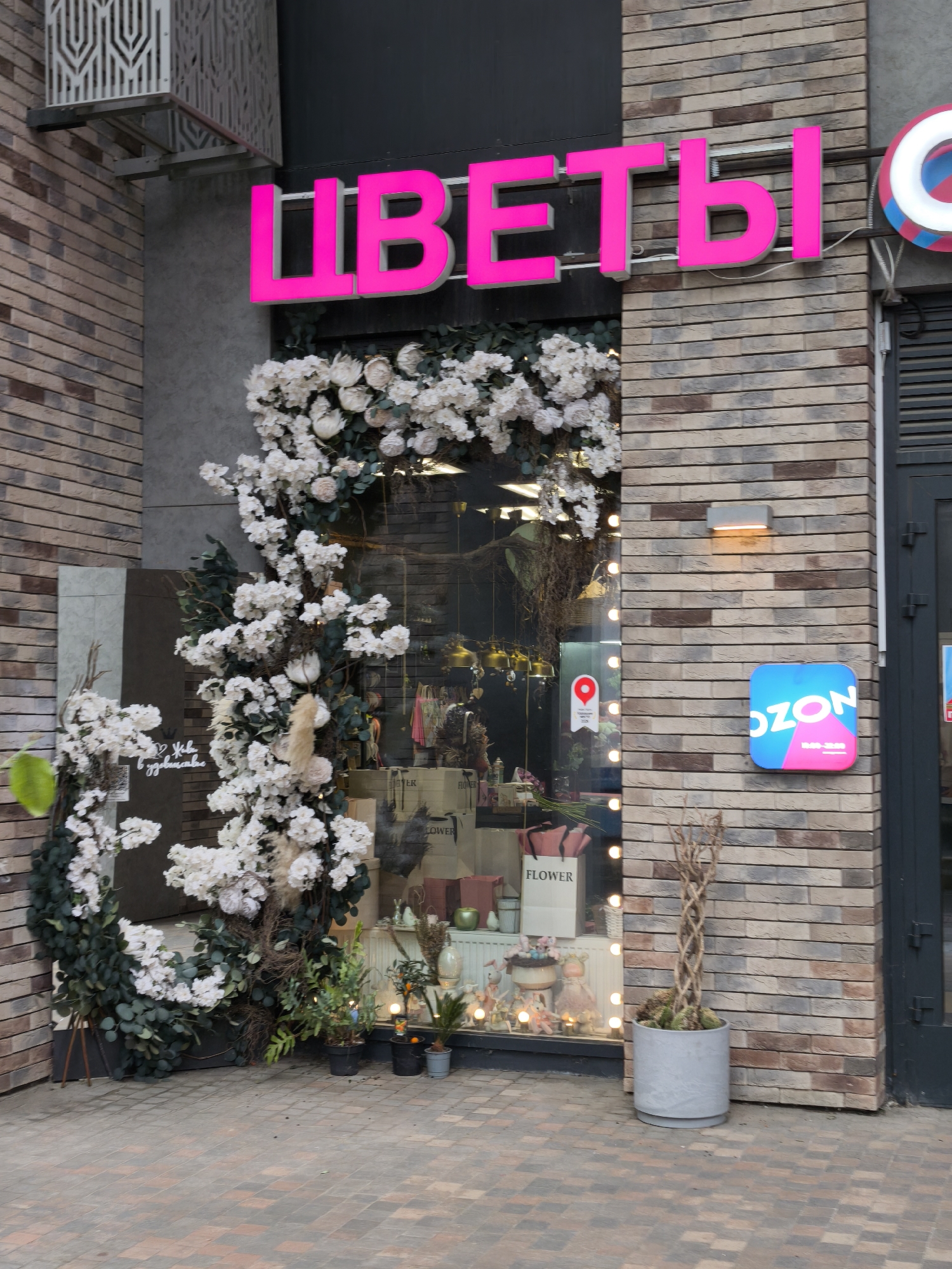 The image shows the entrance to a flower shop in Moscow, Russia. The shop is decorated with artificial flowers, with a pink sign above the entrance that reads "ЦВЕТЫ" (Flowers). There is an OZON sign on the right side of the entrance, indicating that the location may also serve as a pickup point for OZON orders. The storefront is constructed of brick. There is a gray potted plant on the right. The pavement is made of cobblestone. The shop is decorated with a round wreath of white flowers and foliage. Inside the shop window, various floral arrangements and gift items are displayed.