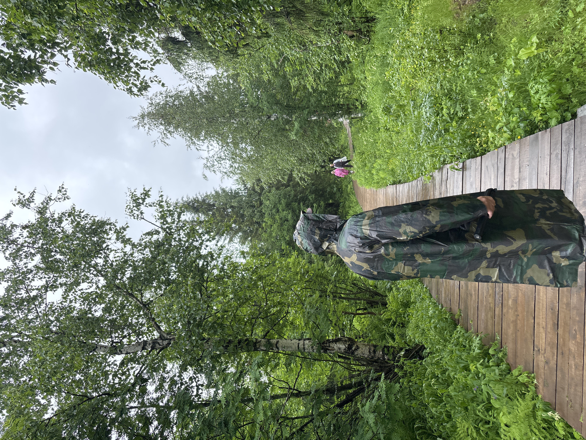 A person wearing a camouflage raincoat walks on a wooden boardwalk through a lush green forest. The boardwalk curves to the right, disappearing into the trees. In the distance, two people in lighter colored clothing are also walking on the boardwalk. The forest is dense with a variety of trees and undergrowth, and the sky is overcast. The scene appears to be from the city of Usva, Russia.