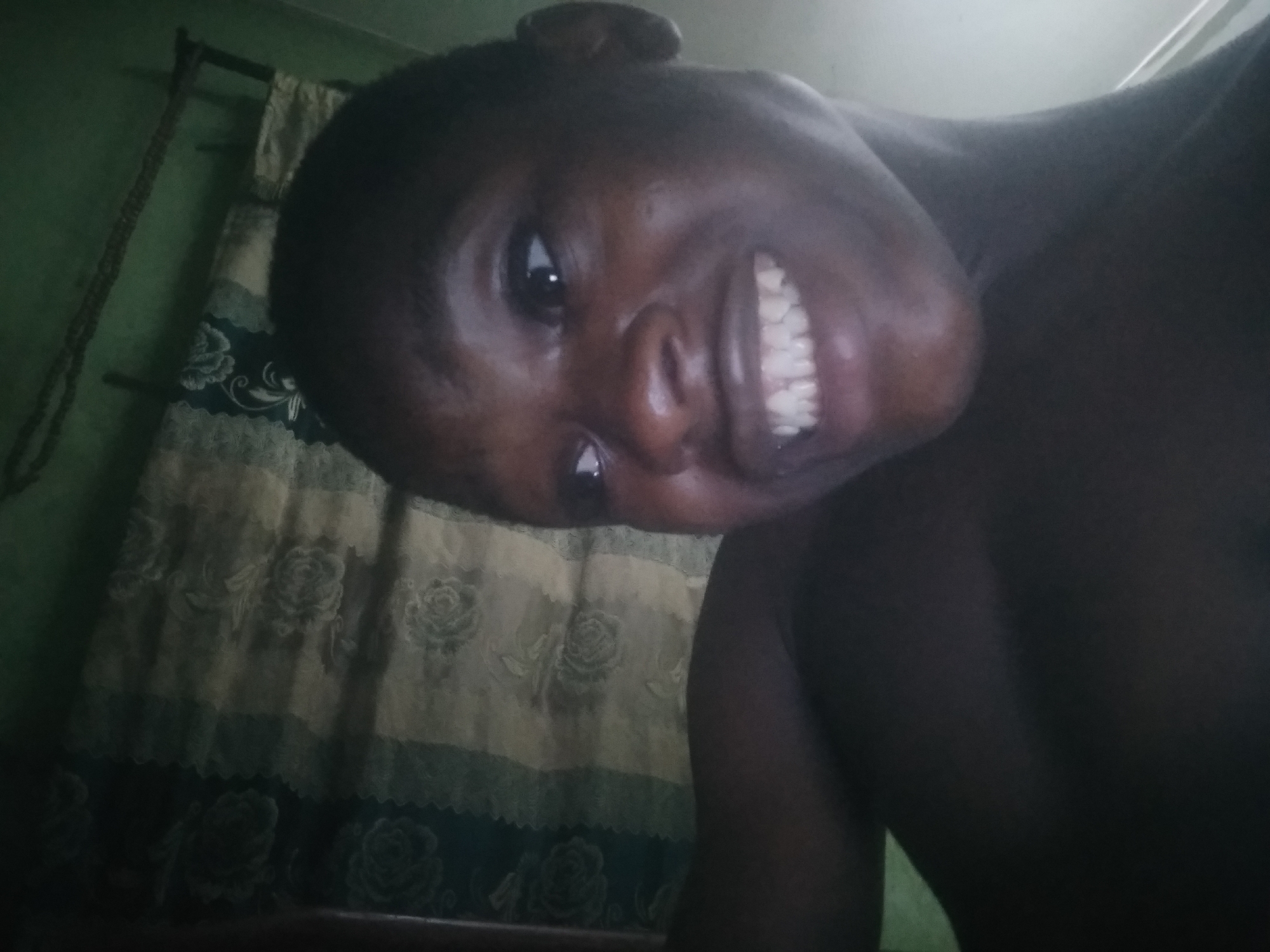 A dark-skinned person with short, dark hair is close to the camera, smiling with their teeth visible. The person's head is tilted to the side. Behind the person is a patterned curtain, predominantly in light and dark shades with a rose design. A green wall is partially visible to the left of the curtain. There's also a dark, rope-like object hanging against the green wall. The location cannot be verified as Shangisha, Nigeria, based on the provided image.