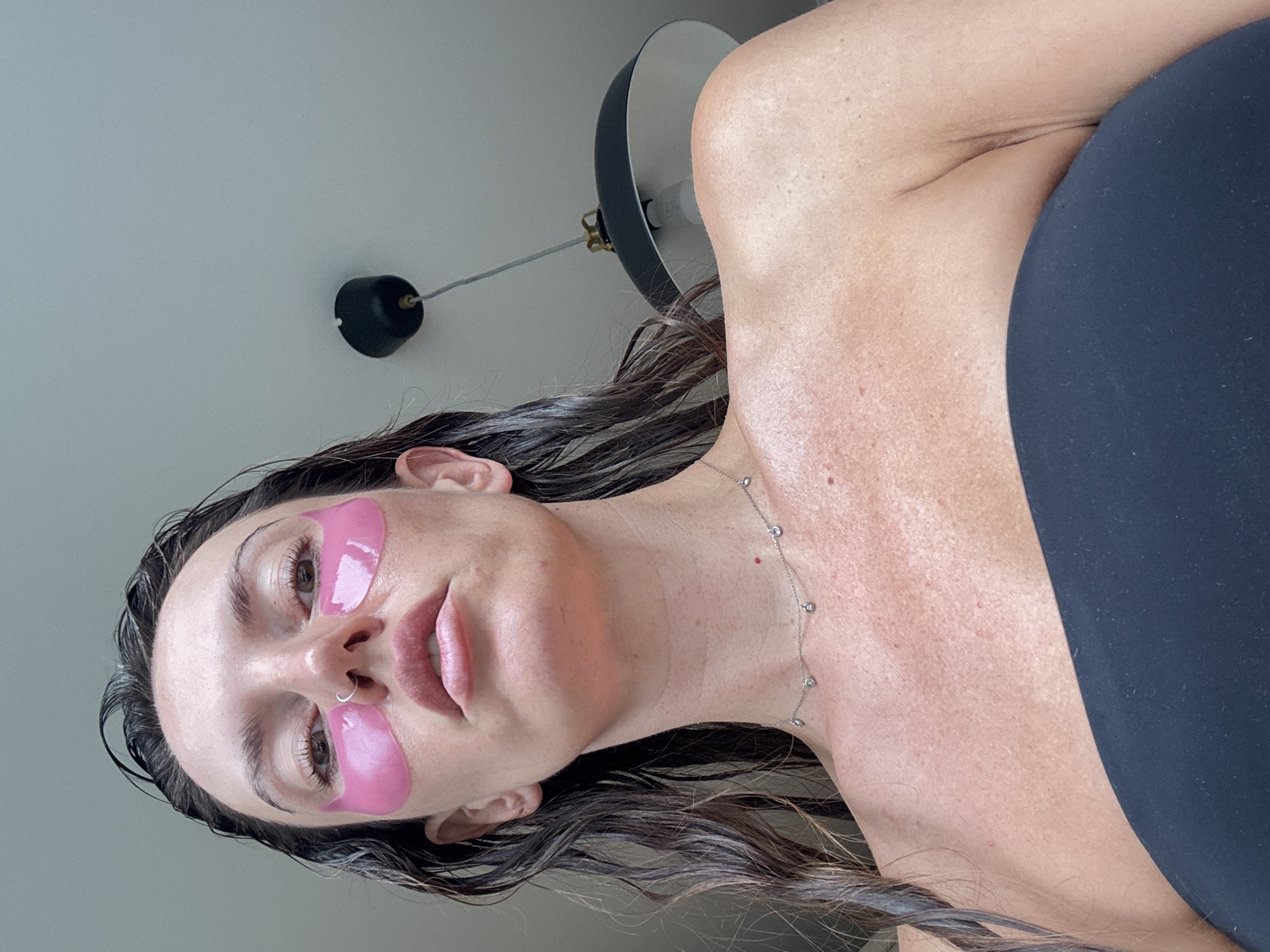 A low-angle, close-up shot shows a woman's face and upper body. She is looking upwards towards the camera, with her head tilted back. She has dark, wet hair that falls around her shoulders. Pink under-eye patches are applied to her skin. She is wearing a strapless black top. A delicate silver necklace with small, clear stones is visible around her neck. The background is a plain, light-colored ceiling with a black hanging lamp suspended from it. There is no indication of the city of Moscow, Russia.