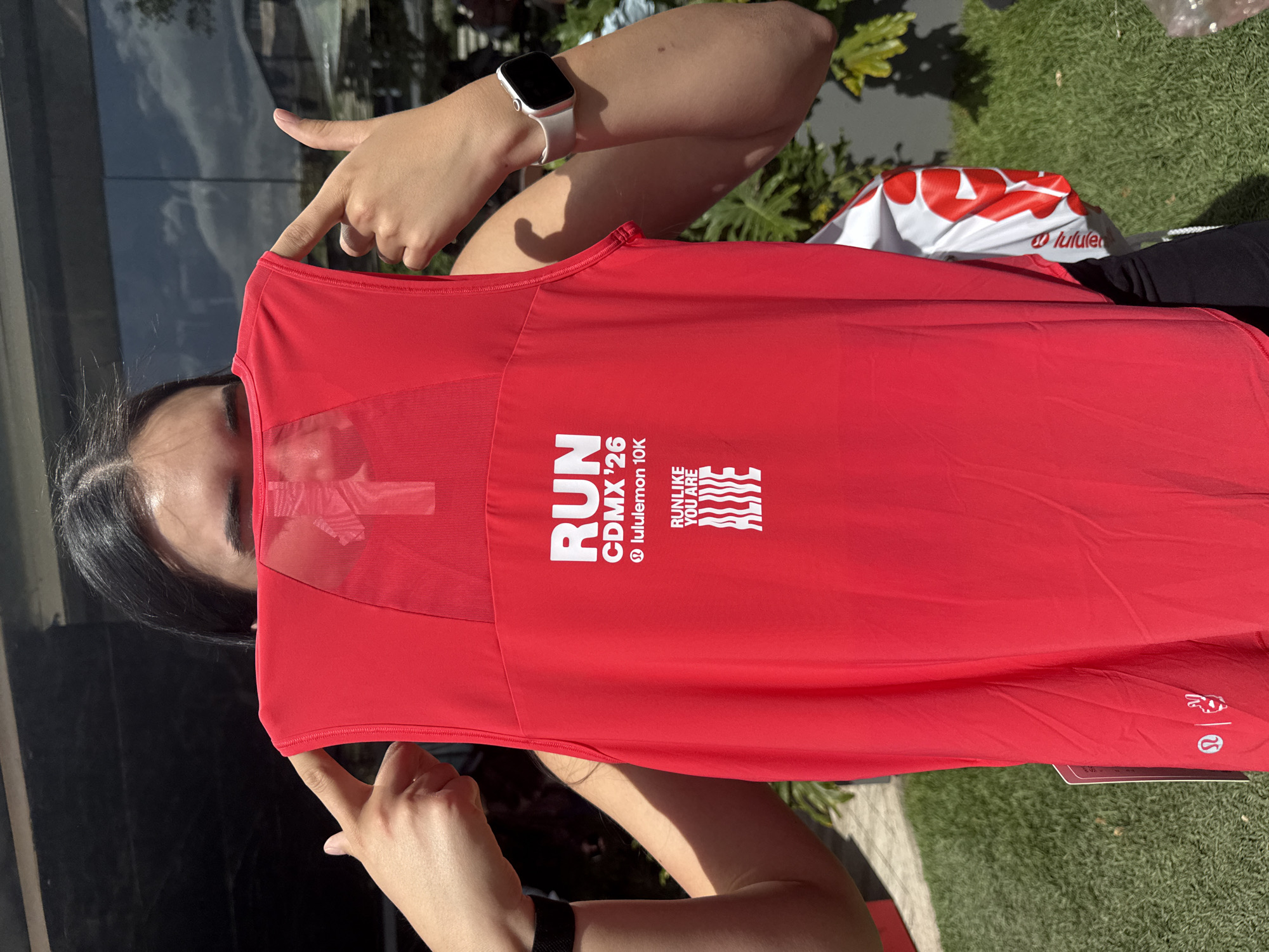 A woman is holding a red sleeveless running shirt that says "RUN CDMX '26 @ lululemon 10K RUN LIKE YOU ARE ALIVE" in front of her face. Her fingers on both hands are pointing at the shirt. She is wearing a white smartwatch. The shirt is a giveaway from the Lululemon race in Mexico City (CDMX). The photo appears to be taken outdoors, possibly near a building with glass windows and some greenery.