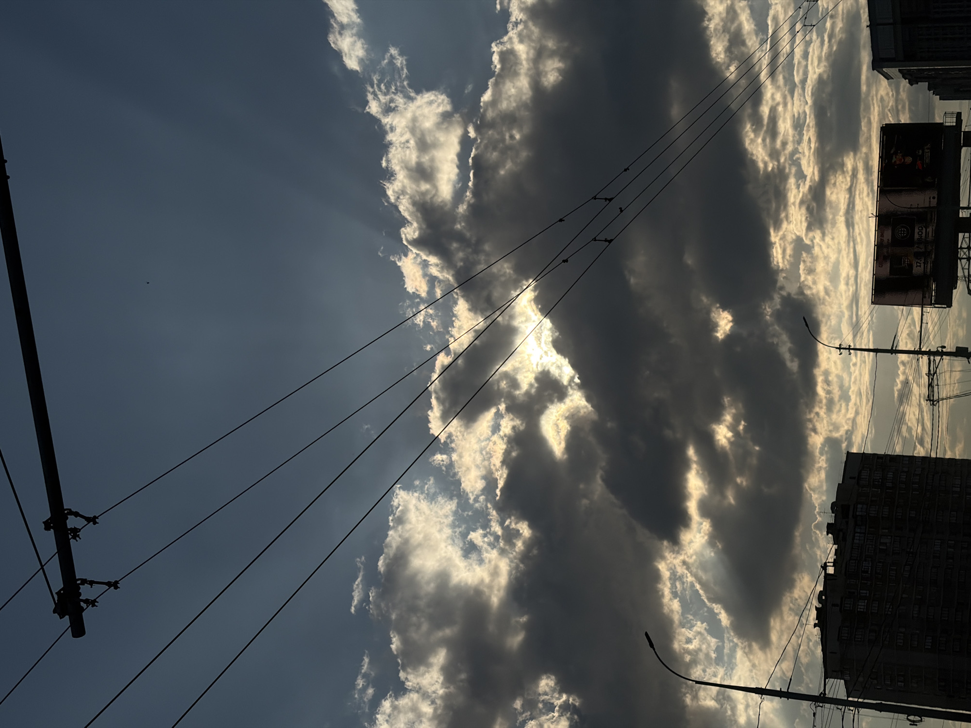 The image depicts a sky filled with clouds, interspersed with patches of clear blue. Sunlight streams through gaps in the cloud cover. Power lines stretch across the frame, originating from a utility pole. The image is oriented such that the horizon line is angled, featuring a building and a billboard structure towards the top. Based on the building style and utility infrastructure, the image can be associated with an urban environment, possibly Krasnodar, Russia.