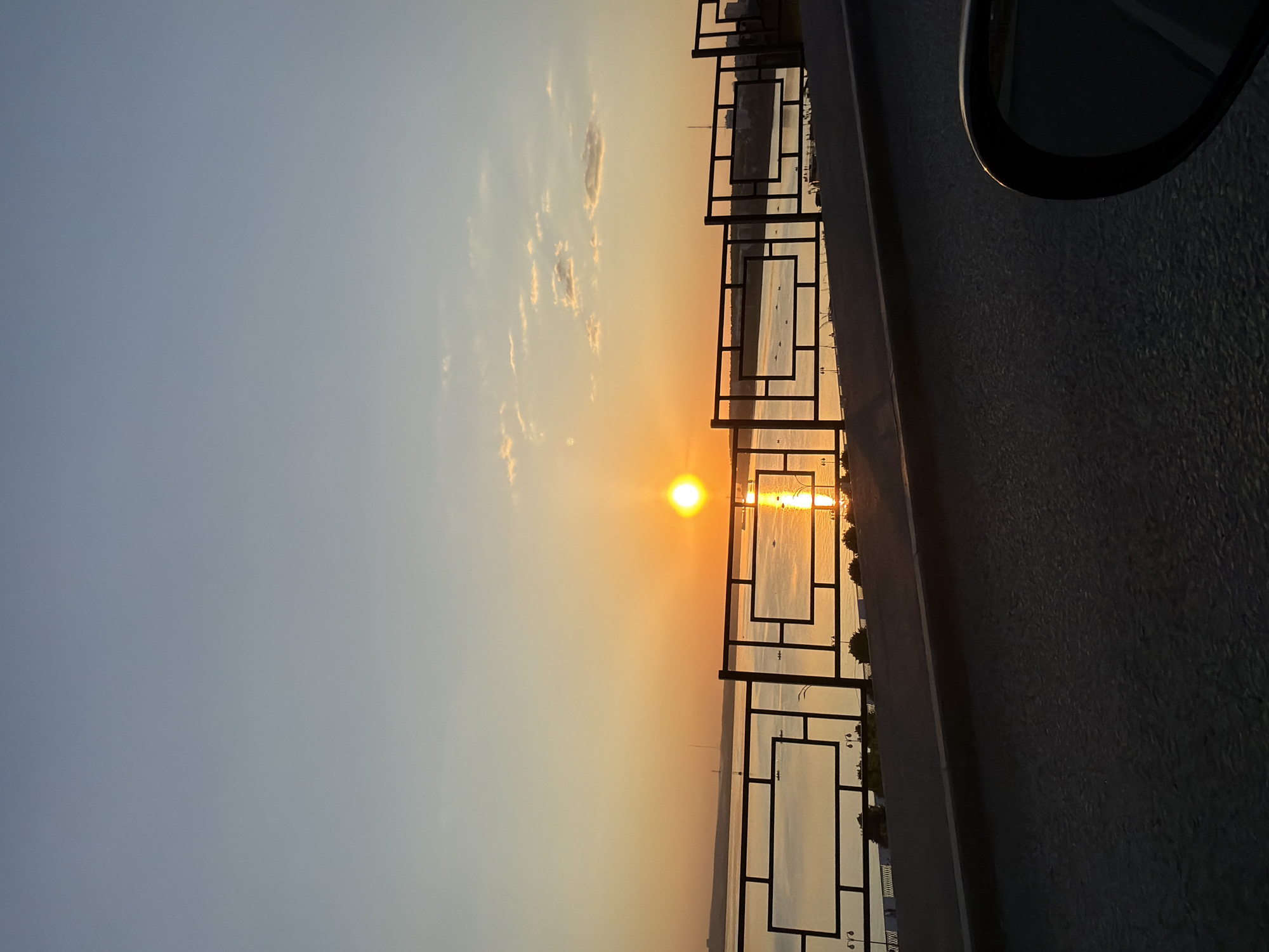 The image shows a sunset view over a body of water, likely a reservoir or large river, with a decorative metal railing in the foreground. The sun is low on the horizon, casting a golden glow across the water and sky. Reflections of the sun are visible on the water's surface. The railing is a modern design with square and rectangular panels. A portion of what appears to be a vehicle's side mirror is visible on the right edge of the image. The sky is a gradient of blue and orange, indicating sunset or sunrise. The location is likely the Izhevsk Reservoir (Izhevsk pond) embankment in Izhevsk, Russia, judging by the railing design and water body. No people are visible in the frame.