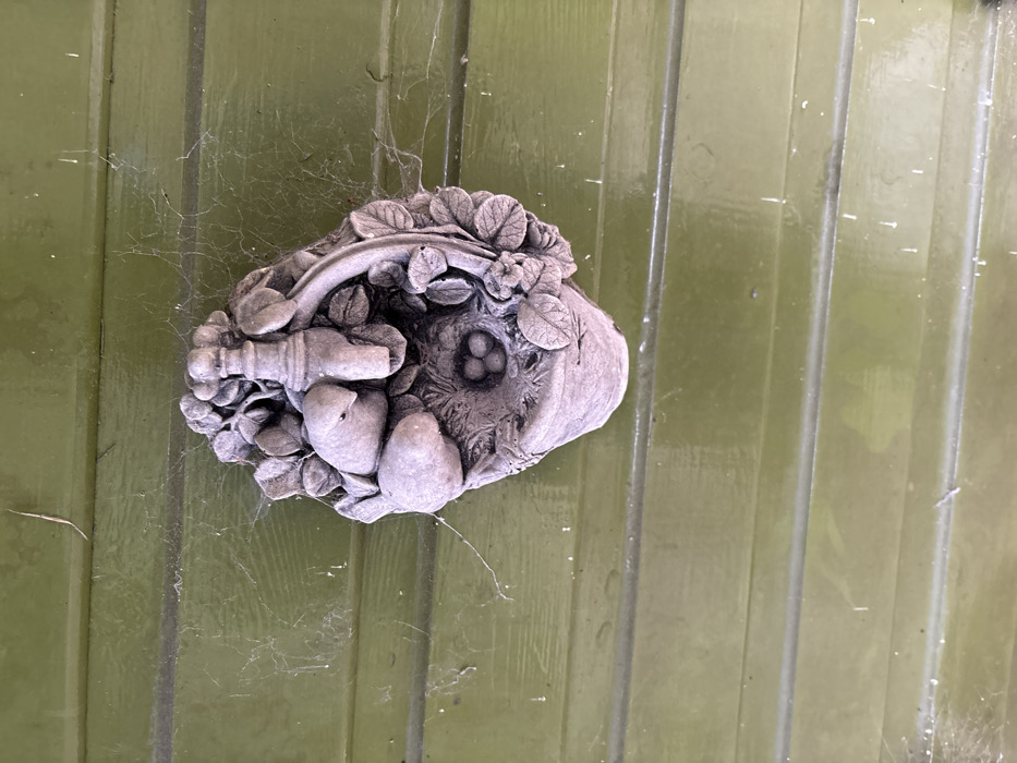 The image displays a weathered, grey, decorative wall ornament mounted on horizontal green wooden paneling in Stiperstones, United Kingdom. The ornament depicts a naturalistic scene, featuring two small, sculpted birds positioned near a nest containing three distinct eggs. Above the birds and nest, a stylized spigot or tap is centrally placed, flanked by floral and leaf motifs that extend around the perimeter, suggesting a basket or planter design. The entire scene is static, with no discernible actions or interactions involving animate subjects. The green wooden paneling exhibits signs of age or exposure, including faded paint and dust accumulation. Multiple delicate spiderwebs are stretched across the green surface, particularly visible to the left and above the ornament, indicating an outdoor or semi-sheltered environment. Small particles of debris are caught within these webs and on the wooden surface.