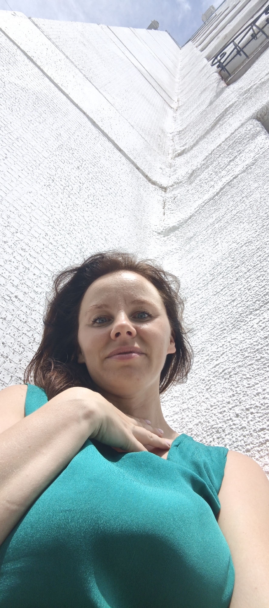 A medium shot, low-angle, outdoor selfie shows a woman with brown hair and blue eyes, looking up at the camera. She is wearing a teal-colored top and has her right hand resting on her chest. Behind her, a textured white wall ascends vertically, meeting the blue sky. The wall appears to be part of a building, with faint lines suggesting brickwork or architectural divisions. A dark, wrought-iron railing is visible in the upper right quadrant, attached to the wall. The scene suggests the woman is standing at the base of a tall building in an urban setting, possibly in Chaikovsky, Russia, given the context.