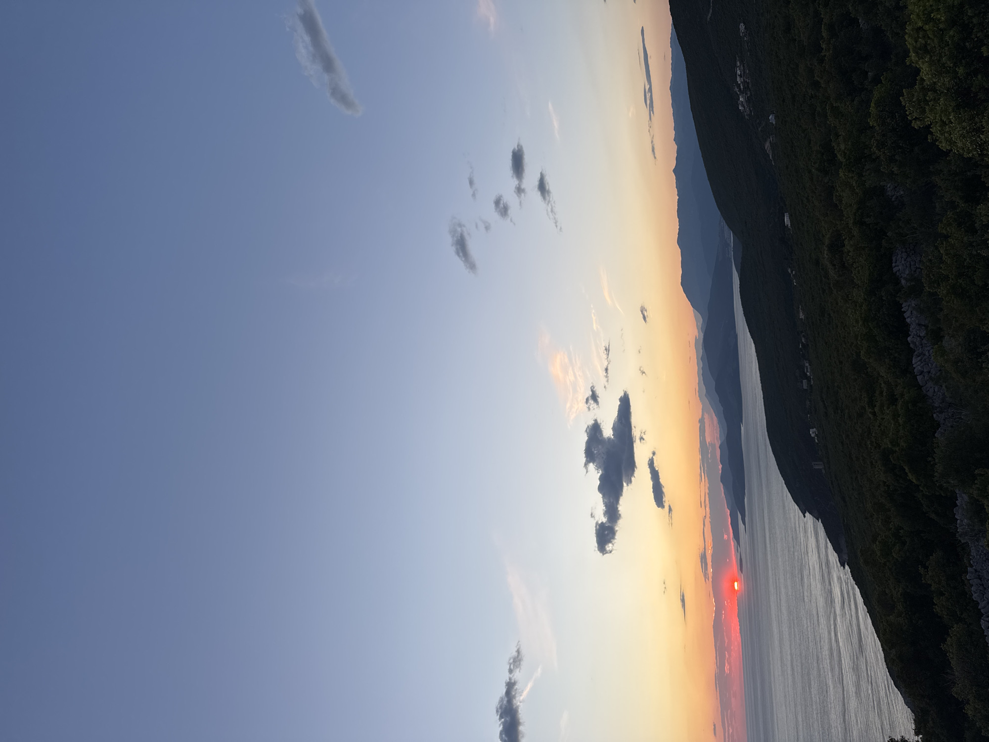 The image shows a sunset over the Adriatic Sea, likely taken from or near Kovačko Polje, Montenegro, given the request in the prompt. The sun, appearing as a bright red orb, is setting on the horizon, reflecting intensely on the water. The sky transitions from blue to yellow and orange as the sun dips lower. Sparse, dark clouds are scattered across the sky. The terrain on the right side is dominated by a steep, green-covered mountain slope, and some outlines of other mountains can be seen in the distance.
