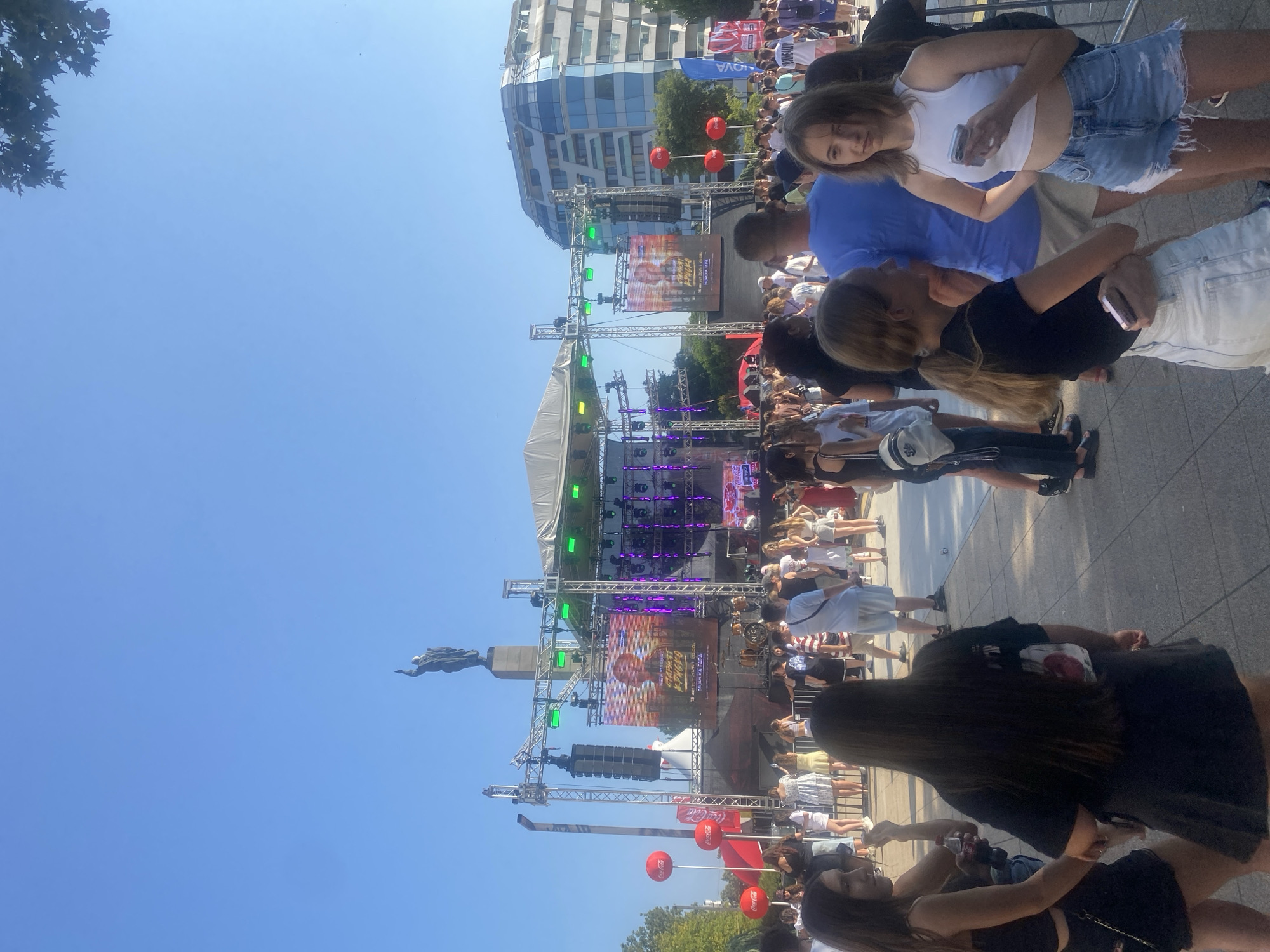 The image depicts a bustling outdoor event, likely a concert or festival, on a clear, sunny day. In the background, a large stage is set up with lighting and screens, and a prominent statue stands atop a pedestal. Several red Coca-Cola balloons are visible, suggesting a sponsorship. A modern, glass-covered building with an undulating facade is situated to the right of the stage. The foreground is filled with a diverse crowd of people, predominantly young adults, dressed in casual summer attire. Many are looking towards the stage or interacting with each other. The event appears to be taking place in the city of Burgas, Bulgaria, as indicated by the background architecture and the statue, which is likely the "Alyosha" monument, a well-known landmark in Burgas. The atmosphere seems festive and energetic.