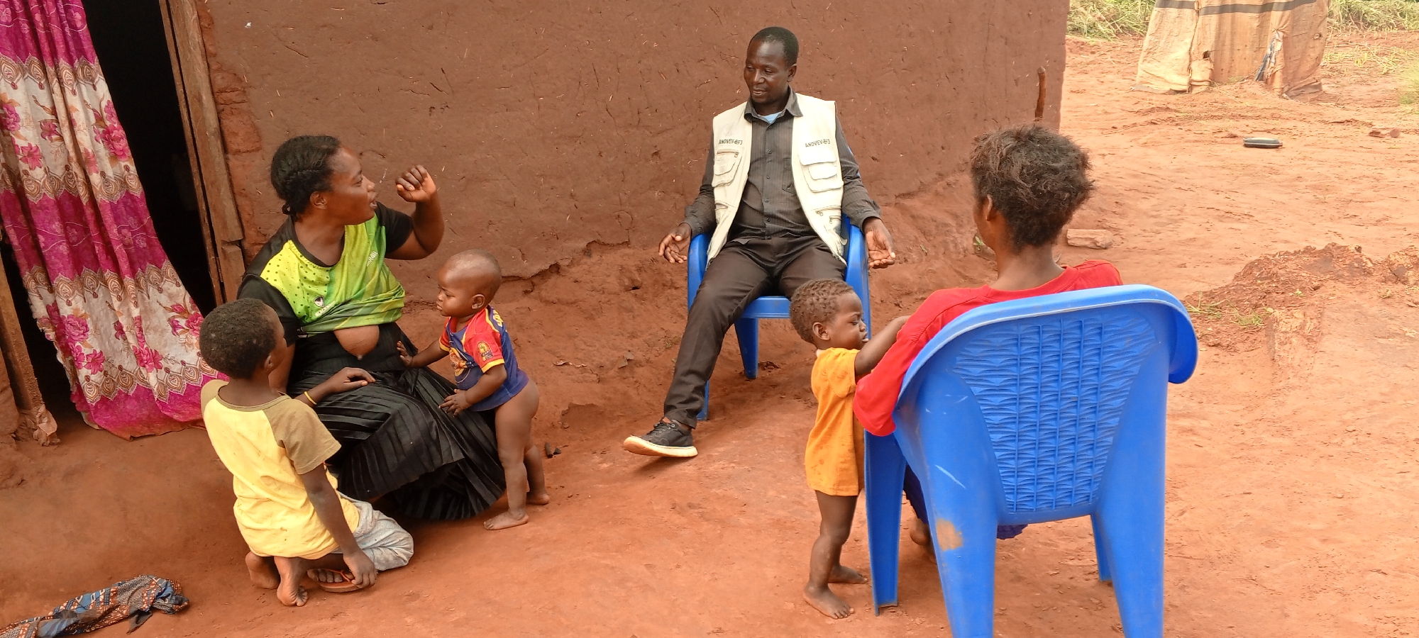 The image depicts an outdoor scene in Makungu, Democratic Republic of the Congo. A man in a vest labeled "ANDVEF" is seated in a blue plastic chair, facing left. To his left, a woman in a green and black shirt sits on the ground with two young children nearby. One of these children is wearing a yellow shirt, while the other is wearing a blue and red shirt. To the right of the man, another woman in a red shirt is seated in a blue plastic chair, facing away from the camera. A young child in an orange shirt stands beside her. The ground is reddish-brown dirt. A building with a pink curtain is visible to the left, and a simple structure made of tarps or fabric is in the background on the right.
