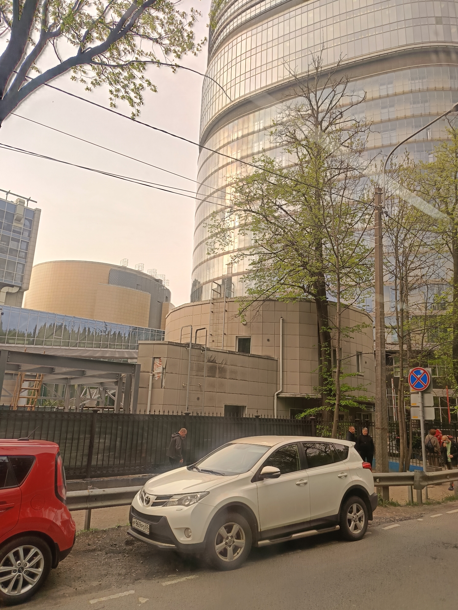 The image depicts a street scene in Saint Petersburg, Russia, likely near a modern architectural complex. Visible buildings include a cylindrical glass structure, potentially an office building, and adjacent structures with beige facades. Two parked cars, a red Kia Soul and a white Toyota RAV4 with license plate P969PO 69, are parked along the curb. A fence runs along the sidewalk, behind which are several pedestrians. A No Stopping sign is visible on the right side of the frame. The sky appears overcast, and trees with fresh spring foliage are visible.