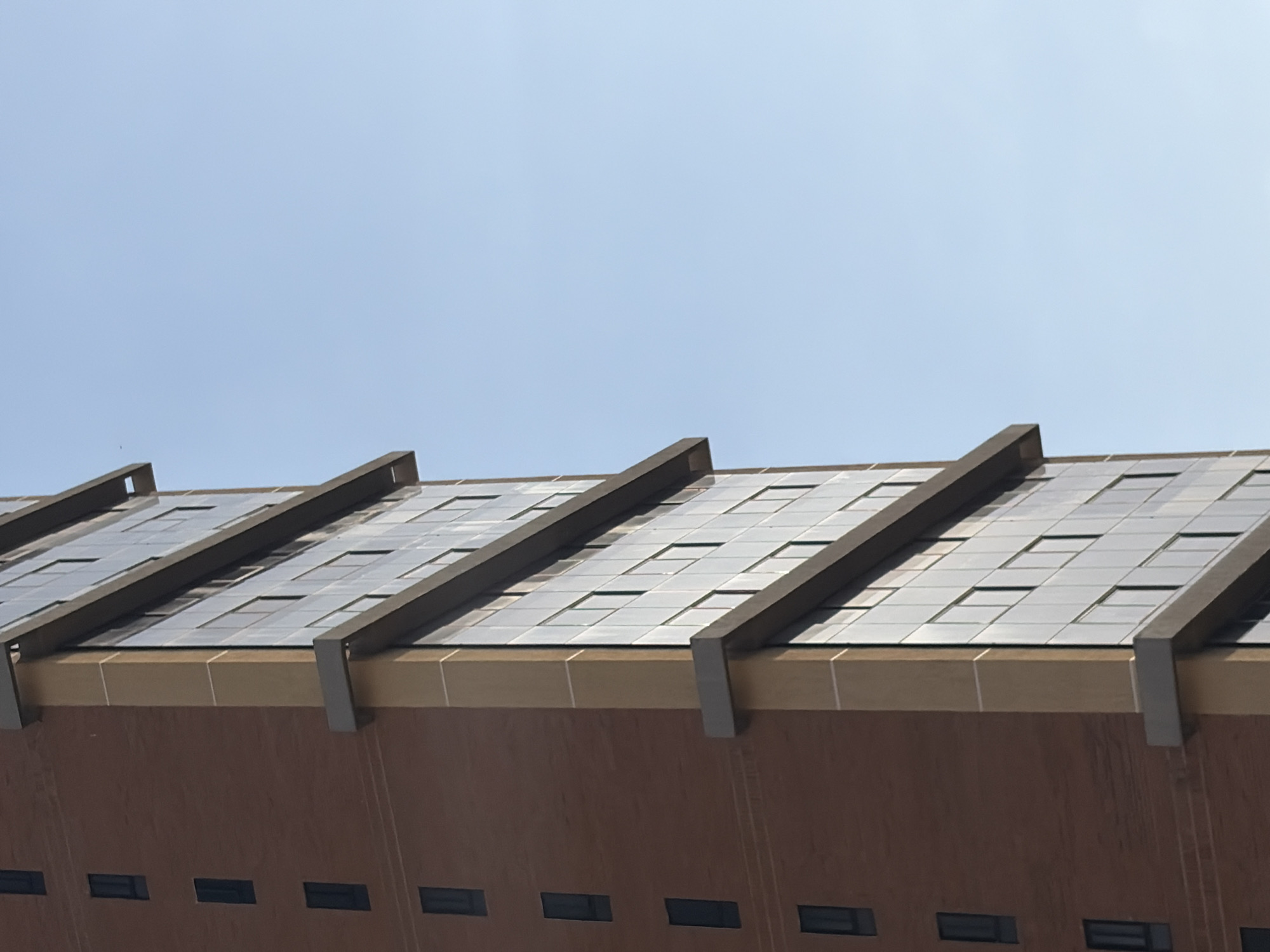 The image depicts the upper portion of a brick building against a clear, blue sky. The building's roof features a grid-like pattern of square tiles, possibly solar panels, aligned beneath a series of parallel, dark-colored, overhanging beams. The brick facade below the roof displays a row of evenly spaced, rectangular windows.
No information about Ciudad Guayana, Venezuela can be extracted from this image. No people, actions, or interactions are depicted.