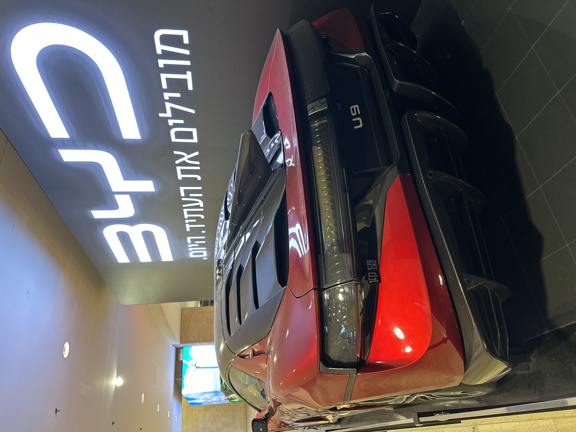 The image shows a red U9 car on display in what appears to be a showroom or promotional space. The text above the car is in Hebrew, and the car is oriented to the right. The scene is located in the city of Tel Aviv-Yafo, Israel. The environment is a modern, well-lit interior space.