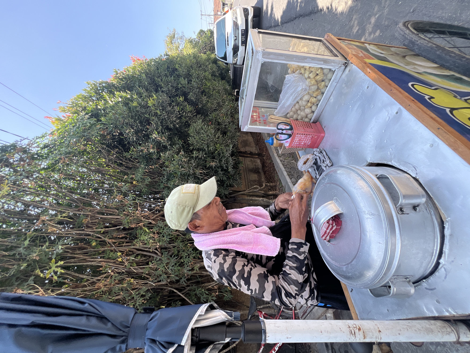 In the city of Denpasar, Indonesia, a man wearing a camouflage long-sleeved shirt, a light green cap, and a pink towel around his neck is preparing food at a street cart. The cart is positioned beside a road with a white SUV parked on the side. The cart has a large, silver steaming pot and a glass display case filled with what appear to be meatballs or similar food items. Various utensils, including scissors and chopsticks, are visible in a pink basket on the cart. Trees and foliage line the background, and the sky is clear and blue. The man is shown in the process of handling a piece of food, possibly wrapping it or adding a garnish. The overall scene depicts a typical street food vendor operation in a bustling urban environment.