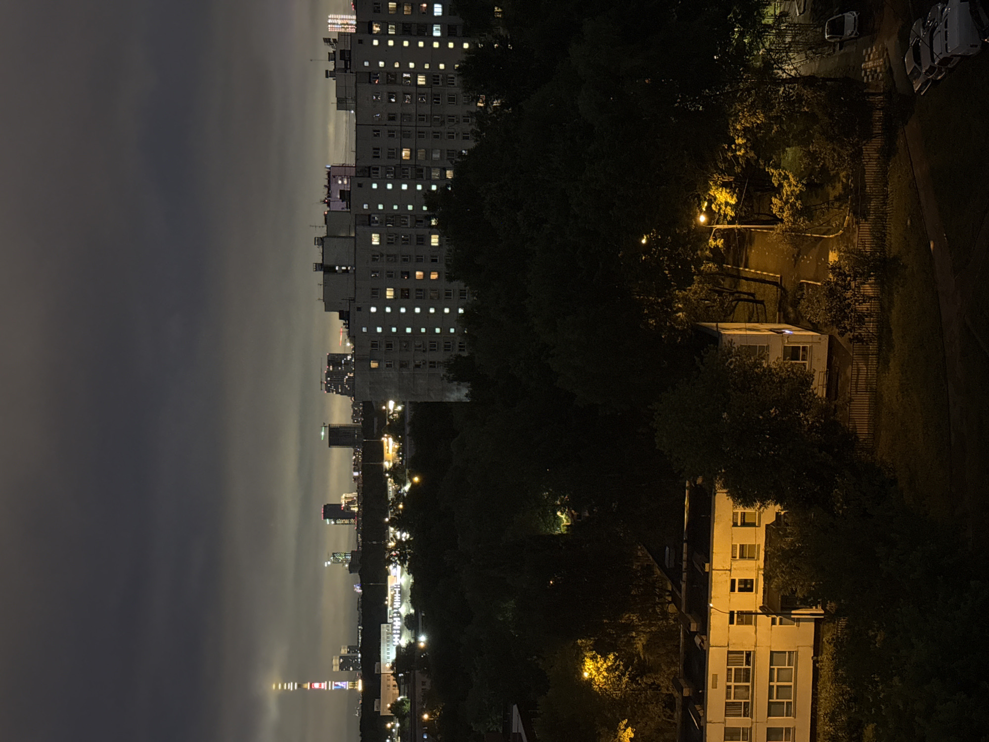 This is an elevated nighttime photograph of a residential area in Moscow, Russia. In the distance, a brightly lit television tower, likely Ostankino Tower, stands tall, surrounded by various other illuminated skyscrapers and buildings forming a city skyline under a dark, cloudy sky.
Closer to the foreground, several large, multi-story apartment buildings are visible, characterized by numerous lit windows, indicating occupancy. One building in the mid-ground appears to be a school or a similar institutional structure, illuminated by yellow outdoor lighting. Dense trees and foliage dominate the area between the buildings, creating dark, shadowy patches. Streetlights cast pools of yellow light onto paved pathways and a road where a single white car is parked. In the lower right corner of the image, several cars are parked on a paved surface. The overall scene depicts a typical urban residential landscape at night, with artificial lights contrasting against the darkness and the natural elements of trees and sky.