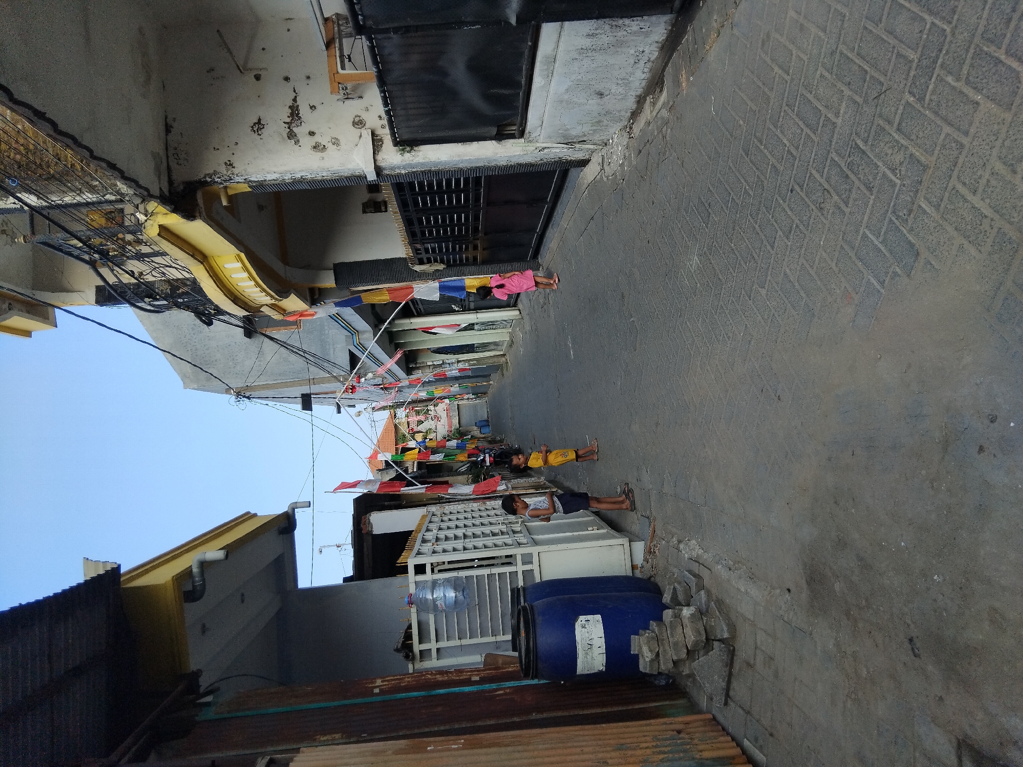 The image depicts a narrow, paved alleyway in what appears to be a residential area of Kepuh Kiriman, Indonesia, likely decorated with Indonesian flags and pennants for a celebration. On the left side of the alley, two young children are standing near two large blue barrels and a stack of bricks. One child, a boy, is wearing a white tank top and dark shorts, while the other, a girl, is in a yellow dress. Further down the alley, another young girl in a pink dress stands facing away from the camera, looking towards a dark metal gate. Buildings line both sides of the alley, with various architectural styles and states of repair visible. The sky is clear and blue. The scene suggests a casual, everyday moment in a densely populated urban environment, possibly during a national holiday or event.