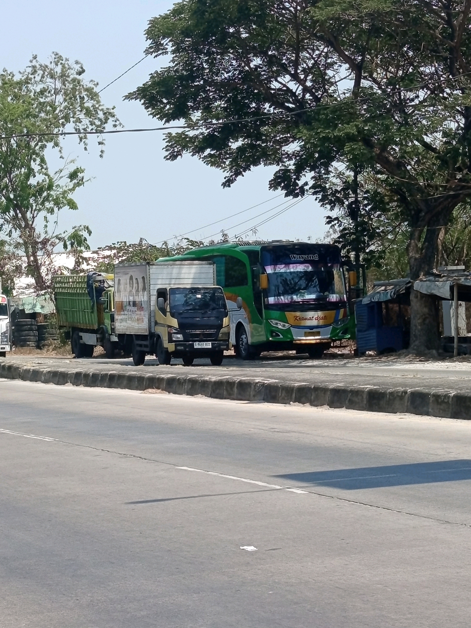 In Karawang, Indonesia, a sunny day showcases a roadside scene with two trucks and a bus parked on the side of a paved road. The foreground features a grey asphalt road with a white line marking its center, and a shadow cast by an unseen object. The background presents a grey concrete barrier, beyond which sits a yellow and black truck, its cargo area covered by a white box with an advertisement featuring portraits. Next to it, a green truck with a ribbed cargo bed is parked. To the right, a vibrant green bus with the word "Wayang" on its front is parked, displaying text that reads "Kramat Djati". Several trees with green foliage line the area behind the vehicles, and a small, makeshift shelter with a blue structure and a weathered grey roof stands near the bus. The sky is clear and blue.