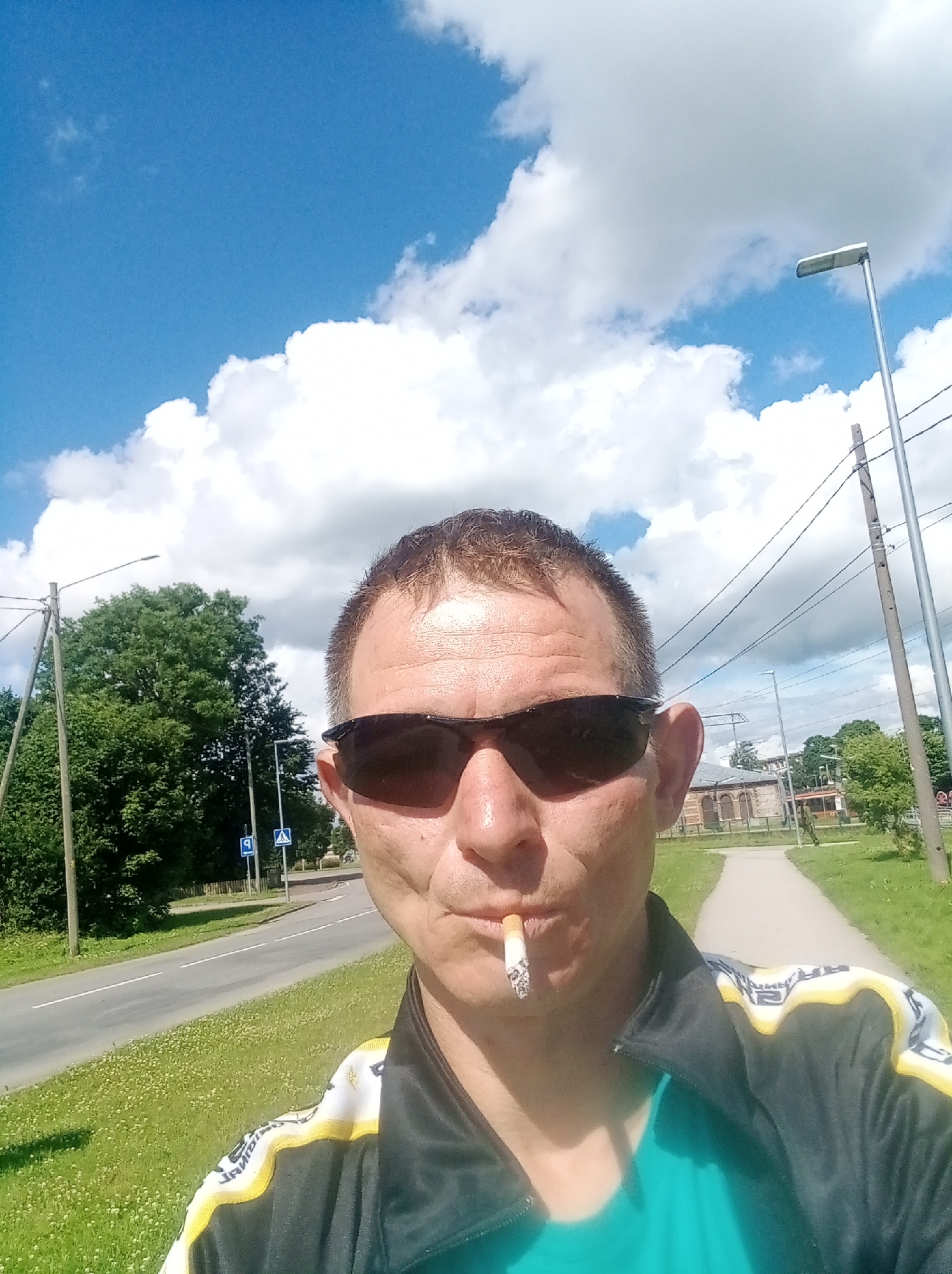 A man with short brown hair and sunglasses is captured in a selfie outdoors. He is smoking a cigarette, which is positioned in his mouth. He is wearing a teal shirt underneath a black jacket with yellow and white detailing on the sleeves. The background reveals a paved walkway and a road, with green grass and trees lining the sides. Two blue and white traffic signs are visible next to the road. In the distance, there are buildings and utility poles with overhead wires. The sky is a bright blue with scattered white clouds, and a streetlamp is visible to the right. The scene appears to be in Tapa, Estonia, based on the provided context.