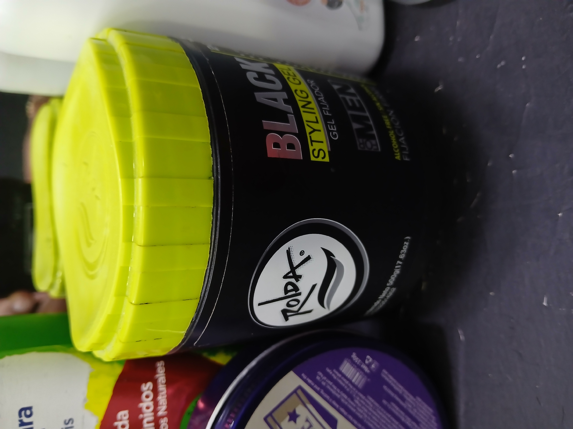 A close-up shot focuses on a container of "BLACK Styling Gel" for men, brand name "ROIDA," with a bright yellow lid. The container is black with red lettering and the text "STYLING GEL," with the text GEL FIJADOR in yellow, and "MEN" in green. The gel is labeled alcohol-free and contains 500g (17.63oz.). The container appears to be sitting on top of other cosmetic or personal care products. The lid of another container is visible in the foreground with purple labeling. Parts of other containers with green and white labeling are visible in the background. The location is not discernable from the image, nor is the presence of people or any actions.