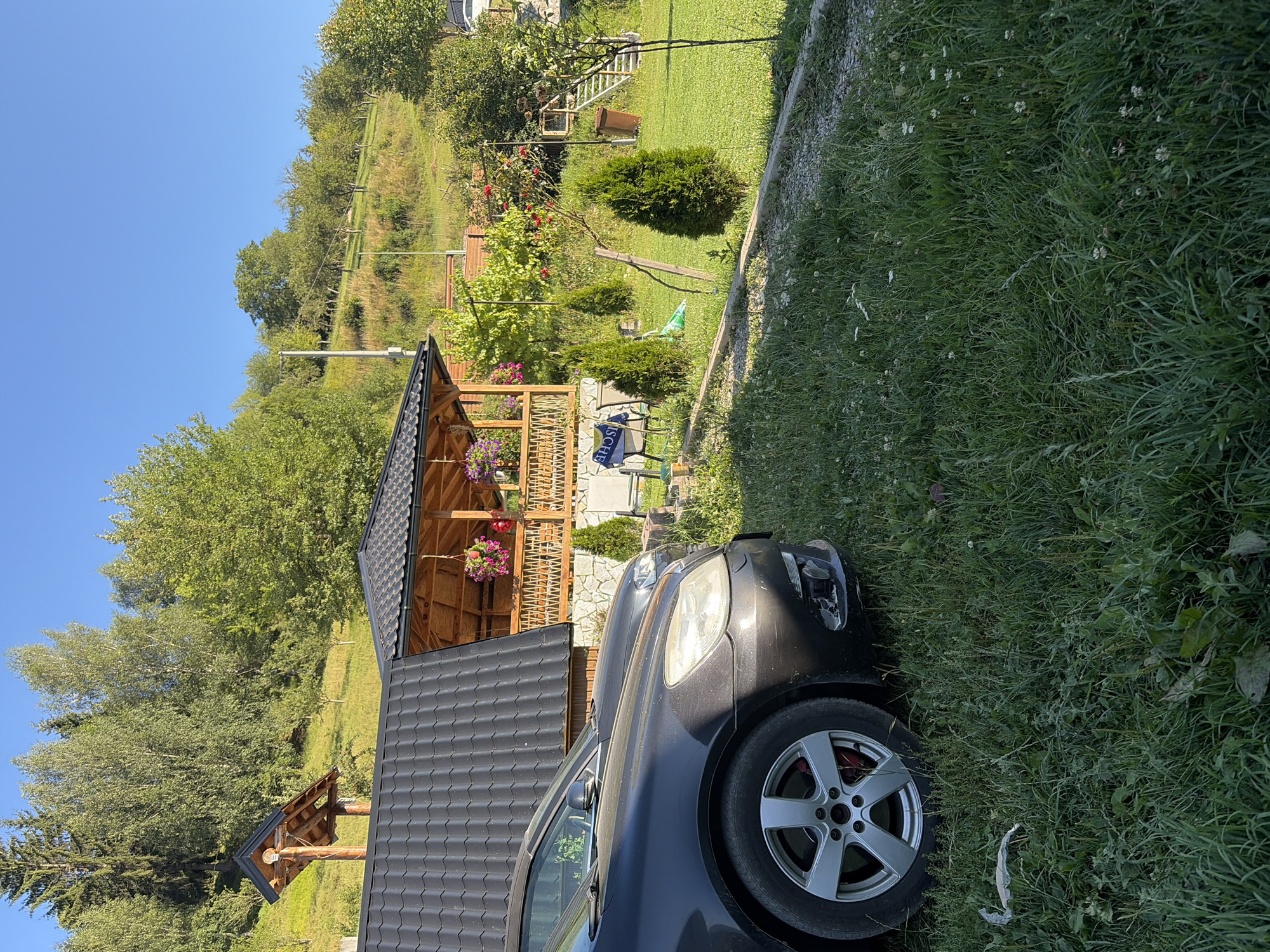 In the city of Plav, Montenegro, a dark gray car is parked on a grassy area in front of a wooden gazebo. The gazebo is decorated with hanging flower baskets and has a railing. Behind the gazebo, a hill covered in trees and vegetation slopes upward. The sky is clear and blue. The scene appears to be a rural or suburban setting on a sunny day.