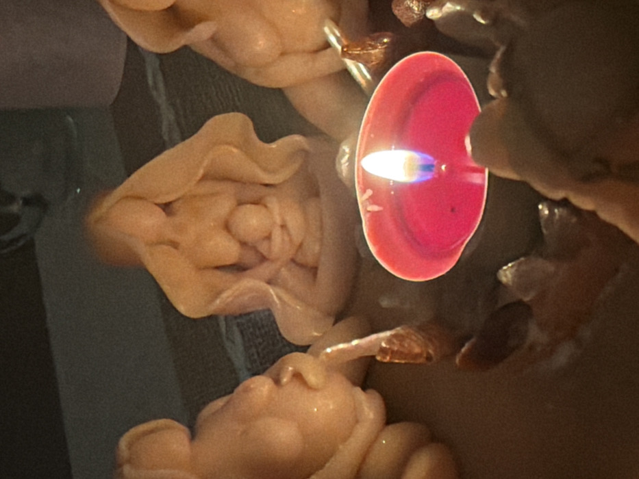 The scene depicts a dimly lit arrangement of wax figures and a burning candle, located in Petah Tikva, Israel. In the immediate foreground, a red votive candle burns with a vertical blue and yellow flame, casting warm light on the surrounding objects. Behind this candle, three peach-toned wax figures are arranged. The central figure depicts a seated female with long, flowing hair cradling an infant close to her chest. To its left, another seated or reclining female figure, visibly pregnant, is partially visible. A third female figure, similar in style, is partially visible to the right. Small, irregular, shiny objects, possibly decorative stones or melted wax remnants, are scattered around the base of the figures and the red candle. The background is dark and out of focus, suggesting an indoor environment. No human subjects are visible or directly interacting with the objects.
