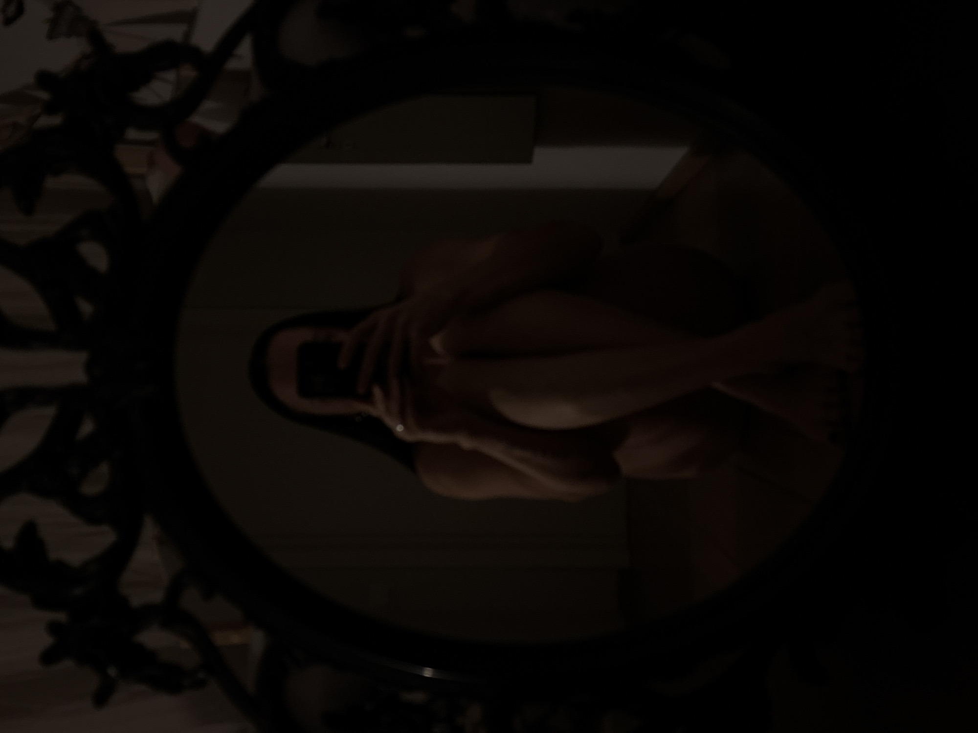 A dark, low-light photograph captures the reflection of a human figure in a circular mirror. The mirror features a prominent, ornate dark frame. Within the reflection, a person is visible, seemingly naked or partially clothed, in a seated or crouching position. Their arms are wrapped around their head or knees, with the head slightly bowed, conveying a sense of introspection, modesty, or vulnerability. The skin tone of the figure appears light brown or beige, standing out slightly against the very dark background of the reflection. The overall scene is extremely dim, suggesting an indoor setting, possibly at night or in a poorly lit room. No specific details of the surrounding room or environment are discernible due to the profound darkness, apart from vague shapes of what might be a wall or floor. There is no visible text in the image. The location context for this image is Chaikovsky, Russia.