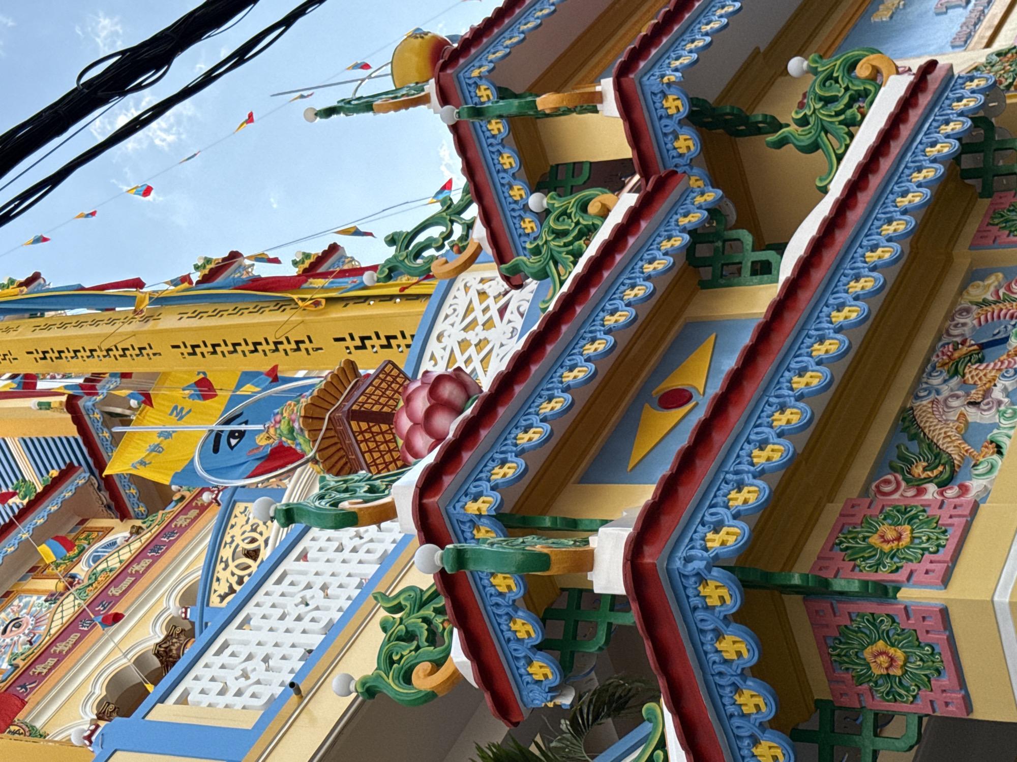 This is a close-up, low-angle, eye-level outdoor shot of a vibrantly colored Buddhist temple in Nha Trang, Vietnam. The building features yellow walls with intricate decorative elements in red, blue, and green. Several levels of ornate roofing are visible, adorned with scrolling green motifs, blue trim with yellow swastika-like symbols, and red eaves. Small, colorful flags in yellow, red, and blue are strung along wires against a bright blue sky with scattered clouds. Power lines are visible in the upper right corner of the image. The overall impression is one of ornate religious architecture, with a focus on detailed decorative patterns and bright, contrasting colors.