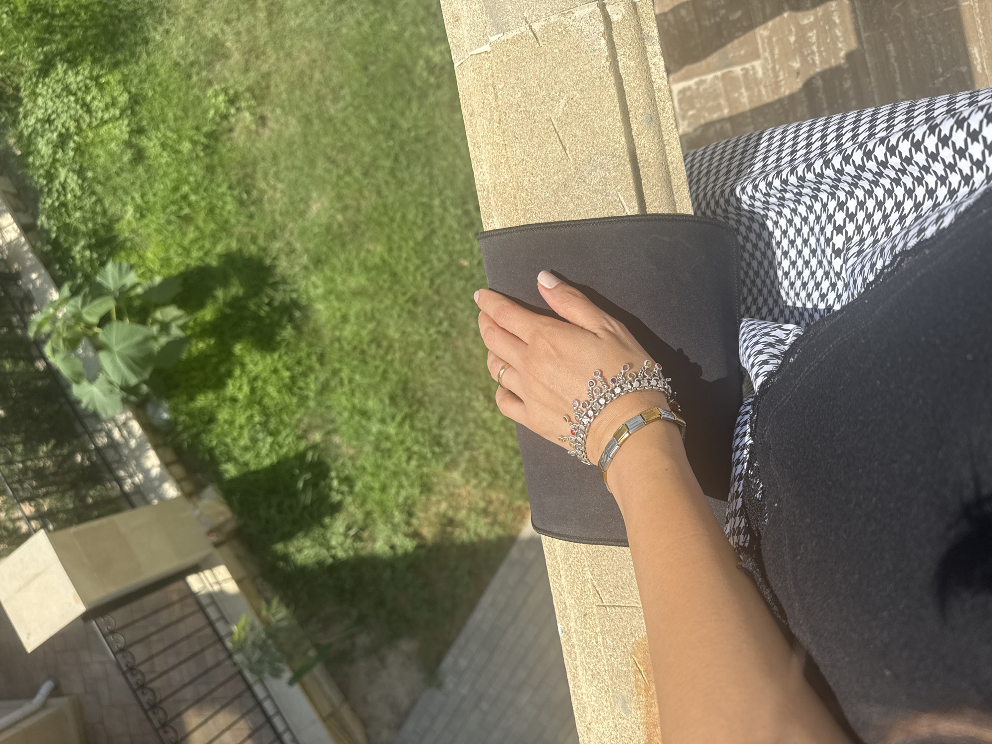 An over-the-shoulder shot captures a woman's arm and hand resting on a dark rectangular object, which is placed on a light-colored, textured stone railing. The woman is wearing two bracelets on her wrist: one is a chain with alternating silver and gold-toned rectangular links, and the other is a more intricate silver-toned bracelet adorned with small clear and pink stones, resembling a crown. A thin gold band is visible on her ring finger. The stone railing extends into the background, with a hint of a black and white houndstooth-patterned fabric beneath the woman's arm. Beyond the railing, a vibrant green lawn is visible, punctuated by a large-leafed plant casting a shadow. A dark metal fence borders the lawn. The scene appears to be outdoors, possibly on a balcony or elevated walkway, in Sabunchu Raion, Azerbaijan, given the architectural style of the railing and the presence of a well-maintained lawn. The lighting suggests a sunny day, with clear shadows indicating a high sun.
