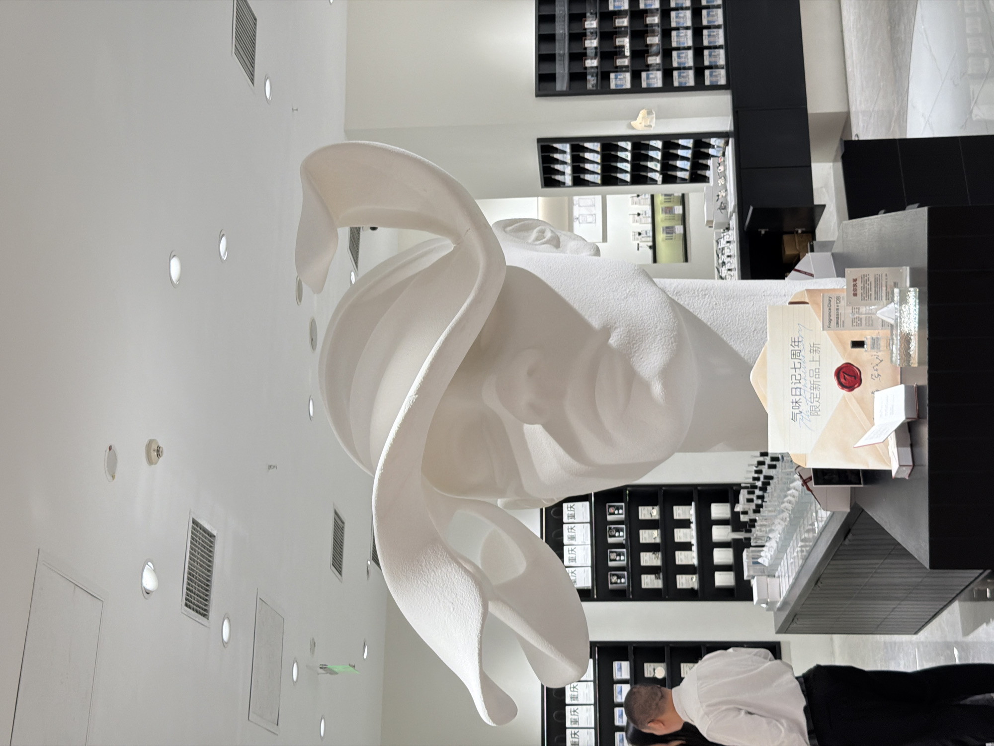 The image depicts the interior of a brightly lit, modern retail establishment located in Yuzhong District, China. The central subject is a colossal, white, matte-textured bust of a human head, possibly female, adorned with a large, undulating, hat-like sculpture. This bust is positioned on a dark, sleek display counter.
To the left, a male individual is visible from the back, wearing a white collared short-sleeved shirt, dark trousers, and dark shoes. He has short dark hair and appears to be observing products on a shelf. A partial view of another person, possibly with dark hair, is barely visible next to him.
Behind the individuals and extending along the walls are multiple black, open-faced shelving units. These shelves are filled with numerous white-boxed products, some arranged in neat rows. Specifically, the top shelves prominently display products featuring the Chinese characters "重庆" (Chóngqìng).
On the dark display counter in front of the bust, various small glass bottles, likely fragrances, are meticulously arranged in rows. A cardboard promotional display board stands prominently, featuring Chinese text that translates to "Fragrance Diary 7th Anniversary" and "New Limited Edition Products," accompanied by a red "7" logo. Nearby, smaller white product boxes, a small fragrance bottle, and a clear acrylic stand with the text "Fragrance Diary" and "重庆日记失落香" (Chóngqìng Rìjì Shīluò Xiāng - Chongqing Diary Lost Fragrance) are displayed.
The ceiling is white and features numerous recessed circular spotlights providing bright illumination, along with several square ventilation grilles, two rectangular access panels, a circular smoke detector, and another small circular fixture. The walls are predominantly white, and the floor appears to be a light-colored, possibly polished concrete or large tile surface.
