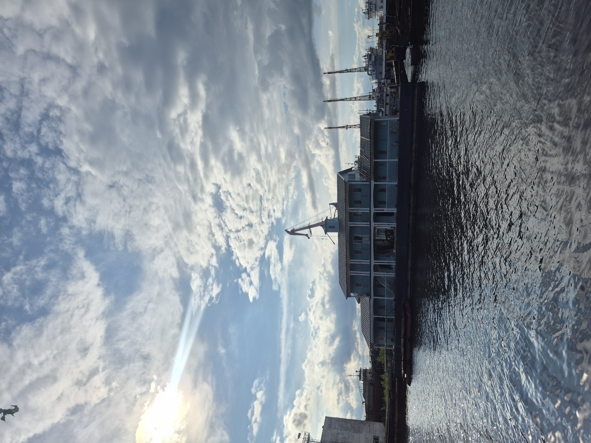 The image depicts a waterfront scene with a view towards the sky. A multi-story, light-blue building is situated along the water's edge. Cranes are visible near the building and further along the waterfront. The sky is filled with clouds, and sunlight is visible in the lower portion of the frame. The water is rippled, reflecting light. The location may be in or near Chaikovsky, Russia, given the query.