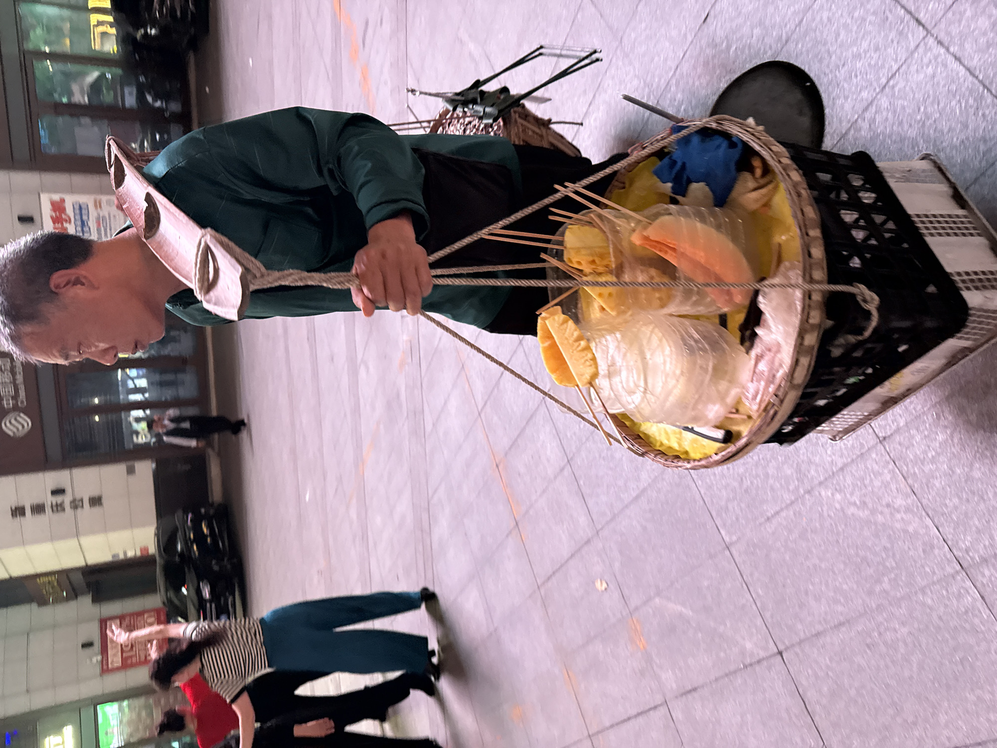 A street vendor carries food on a shoulder yoke in Yuzhong District, China. The vendor, an older person wearing a green shirt and a black apron, supports a woven basket on each end of the yoke. The baskets contain food, some skewered with sticks and packaged in clear plastic. Other items like a blue cloth and metal utensils are also visible. In the background are blurred images of people walking, street lamps, and a building with a sign written in Chinese characters. The ground is paved with light-colored tiles.