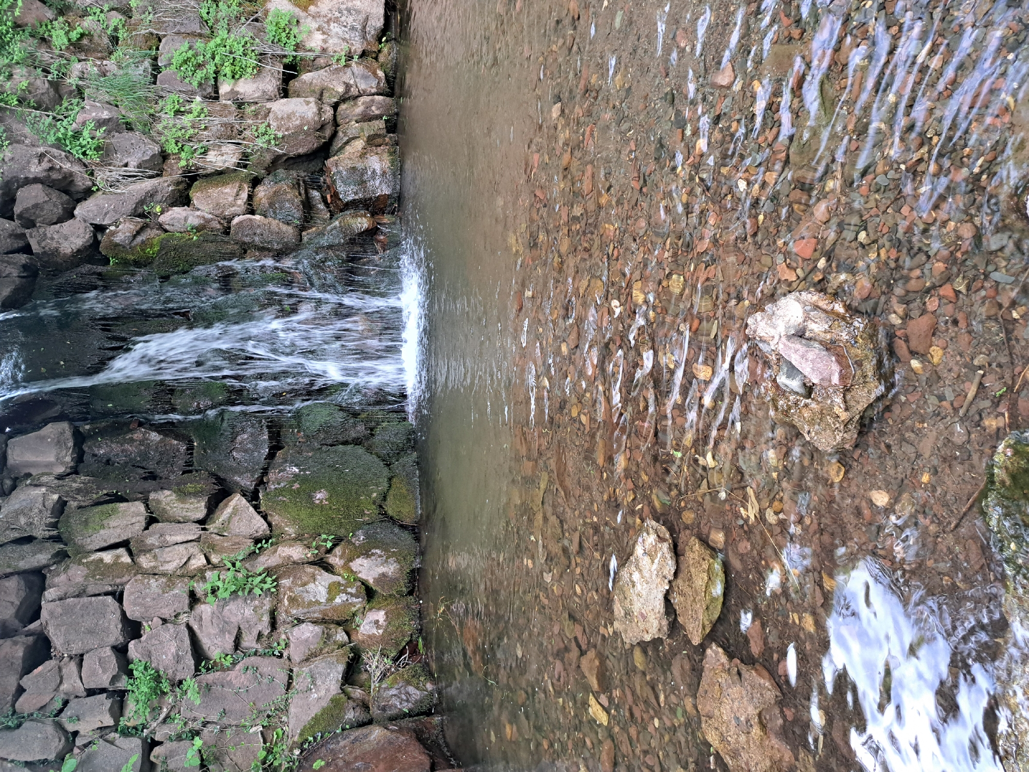 The image depicts a natural waterfall cascading into a shallow, clear body of water in Lokorsko, Bulgaria. The waterfall originates from a rough stone wall that is partially covered in moss and small green plants. The water flowing over the rocks creates a frothy, white curtain, and the pool below is filled with submerged pebbles and stones of various colors and sizes. The water's surface is rippled, reflecting the surrounding environment. Large, rough stones are visible along the edge of the water and within the pool. The overall impression is of a serene, natural setting within the vicinity of Lokorsko.