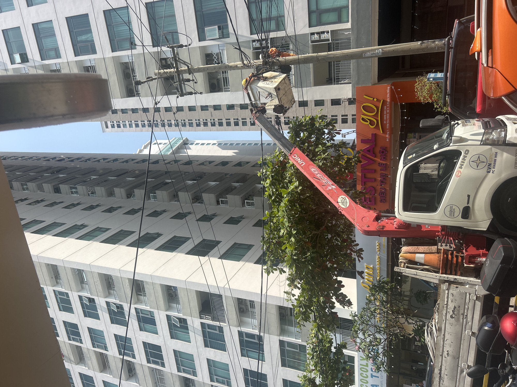 A utility worker on a crane is repairing or maintaining overhead electrical equipment on a utility pole. The crane is mounted on a white HINO truck. The truck has the insignia of EVN CPC and PC KHANH HOA, suggesting the utility company is involved in electricity distribution for the Khánh Hòa province. In the background, there are tall residential or commercial buildings. The location is in Bắc Nha Trang, Vietnam, indicated by the Vietnamese text on the "FESTIVAL go!" sign. The sign also includes a phone number and an address, implying this is a commercial area, possibly near the TTTM Tòa OC2. Other businesses visible are “COCOLY” and “Anna”.