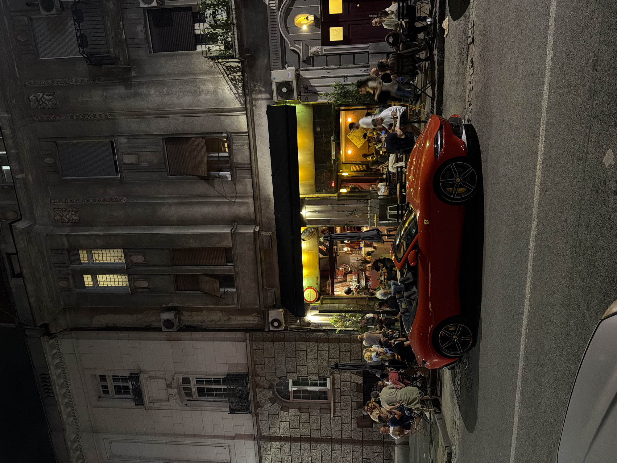 The scene depicts a vibrant nighttime street in the Vracar Urban Municipality of Belgrade, Serbia. An illuminated restaurant spills onto the sidewalk, where patrons sit at tables. A red sports car is parked alongside the restaurant, partially obstructing the sidewalk. Buildings with varying architectural styles flank the street, featuring windows and decorative facades. People are seen eating, drinking, and socializing. The street is paved and features painted lane markings. Another vehicle is parked near the sports car.