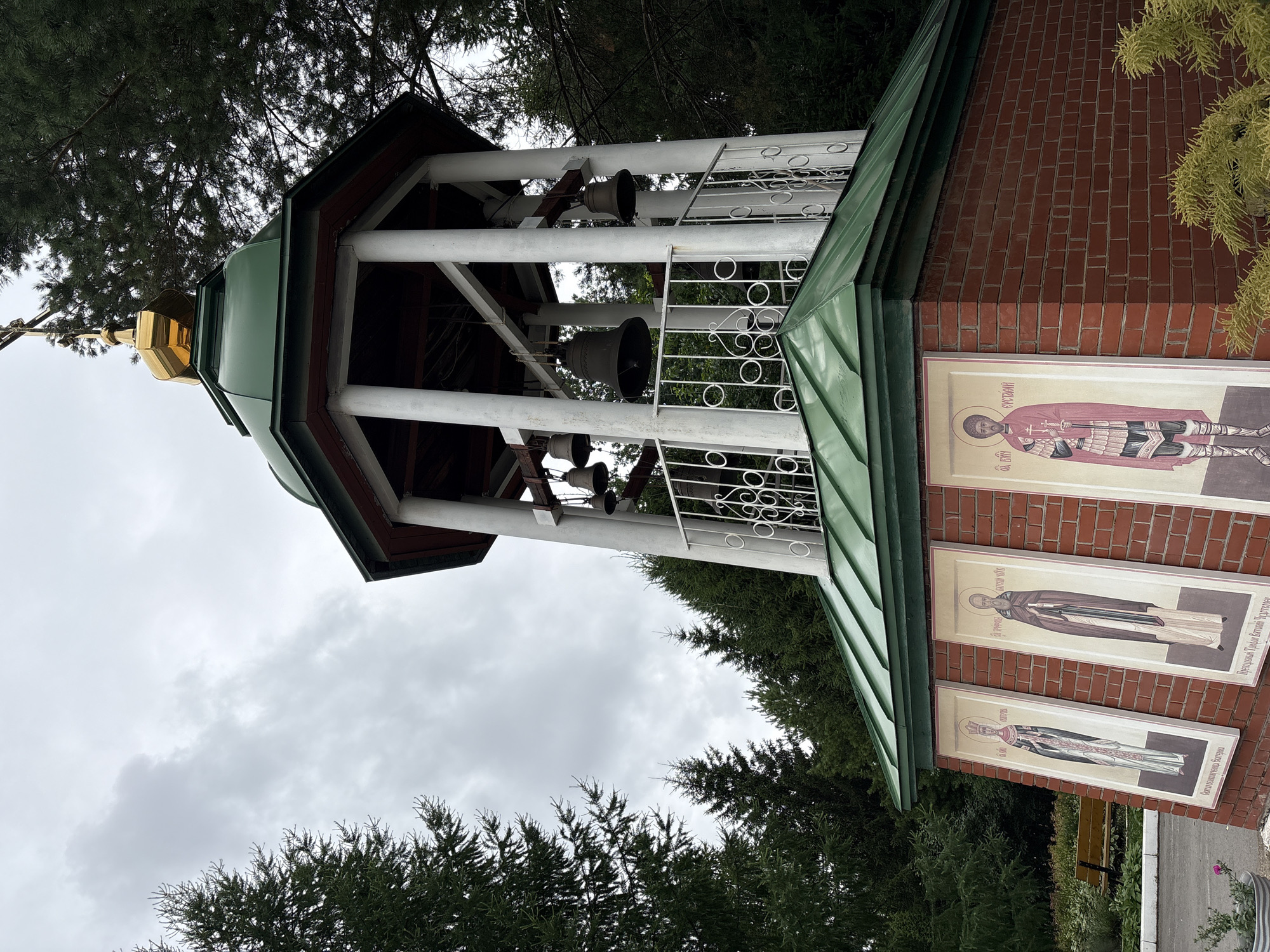 The image shows a bell tower with a green roof and a golden dome at the top, adorned with a cross. The tower is supported by white pillars and has several large bells hanging inside. The base of the tower is made of red brick and features three religious icons painted on vertical panels. The icons depict figures in traditional religious attire. The bell tower is surrounded by tall evergreen trees and is situated under a cloudy sky. In the foreground, there is some green foliage and a bench can be seen to the left of the tower. The scene appears to be a part of a religious complex or a churchyard in Perm, Russia.