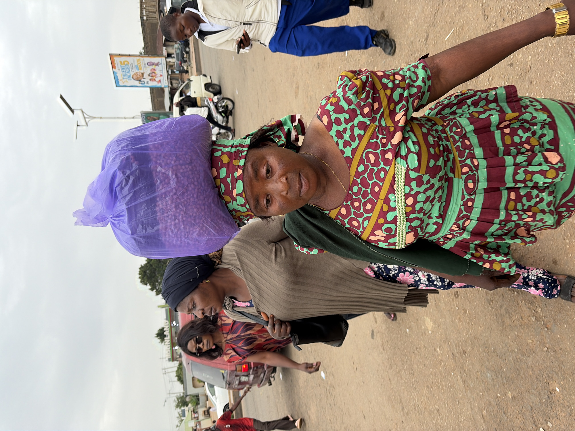 This is a medium shot, eye-level, outdoor photograph taken in Gura Top, Nigeria, depicting a street scene with several individuals. In the foreground, a woman with dark skin wears a vibrantly colored dress with a green and burgundy leopard-like print. She has a large, translucent purple bag filled with small purple items balanced on her head. She also wears a green garment draped over her shoulders and a gold watch on her right wrist. Behind her, another woman, wearing a black headscarf and a long, ribbed brown sweater over a floral-patterned dress, walks with a black purse slung over her shoulder. Further in the background, a person in a red shirt and dark pants walks from left to right. To the right of the foreground woman, a young man in a white jacket and blue pants is looking at his phone. A white van and a red SUV are visible in the background, as well as a billboard with colorful images and text. The ground is dirt and gravel, and the sky is overcast. The overall impression is of a busy, everyday street scene.