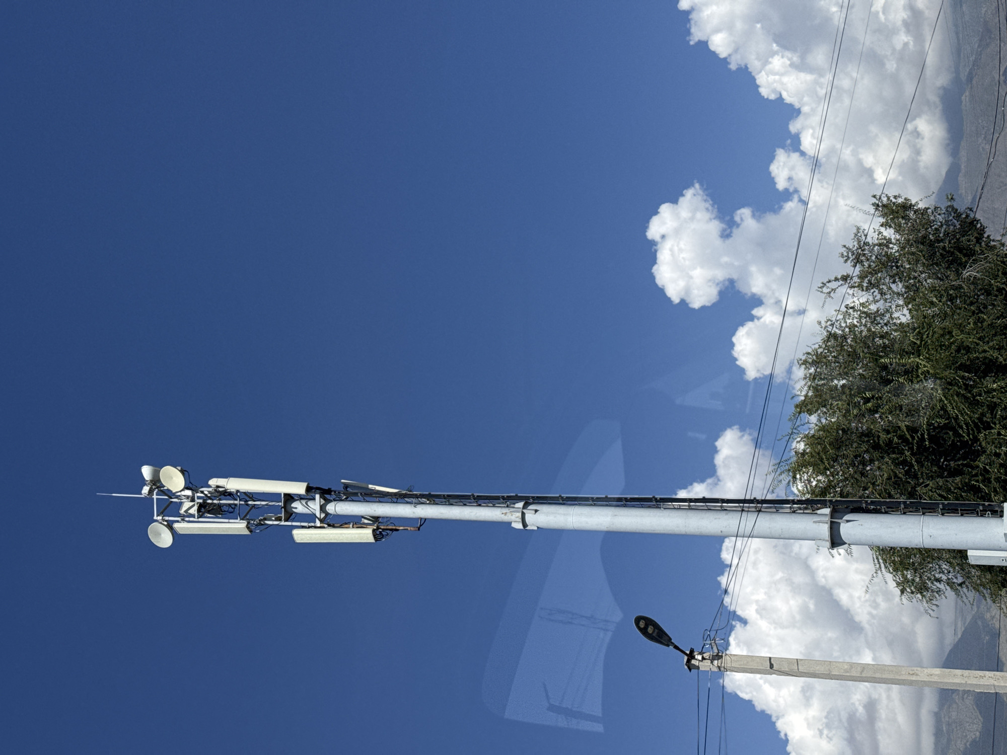 A tall, grey metal communication tower stands against a bright blue sky with fluffy white clouds. The tower is equipped with multiple antennas and dish-shaped receivers at its apex. To the left of the tower, a grey concrete pole supports a black street lamp. A dense, green tree occupies the lower right portion of the frame. In the distance, mountains are faintly visible. The image appears to be taken in Tamchy, Kyrgyzstan, likely on a clear day.
