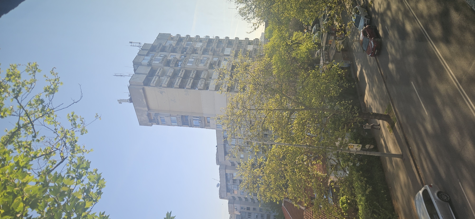 The image captures a sunny daytime street scene in Plovdiv, Bulgaria, likely during spring or early summer, given the vibrant green foliage. The perspective is elevated, looking down onto a street lined with trees and buildings.
Dominating the background is a tall, multi-story, concrete apartment building. It features numerous balconies and external structures, possibly air conditioning units or antennas, suggesting a typical Eastern European post-socialist era residential block. Other, less distinct buildings are visible further along the street.
In the mid-ground, a row of deciduous trees with fresh, bright green leaves runs parallel to the street, partially obscuring the buildings behind them. These trees cast long, dappled shadows across the scene. Below the trees, a paved road with double white lines is visible, on which several cars are parked or moving. A red car and a silver/white car are clearly discernible.
A sidewalk runs alongside the road, where one person can be seen walking. A street pole stands on the sidewalk, featuring a yellow sign with an arrow pointing right and a white circular sign below it. Through the trees, a partially visible sign reads "БЪРЗА ХРАНА" (Bǎrza Khrana), which translates to "Fast Food." The sky above is clear and blue, indicating good weather. The overall impression is of a bustling urban environment on a pleasant day.