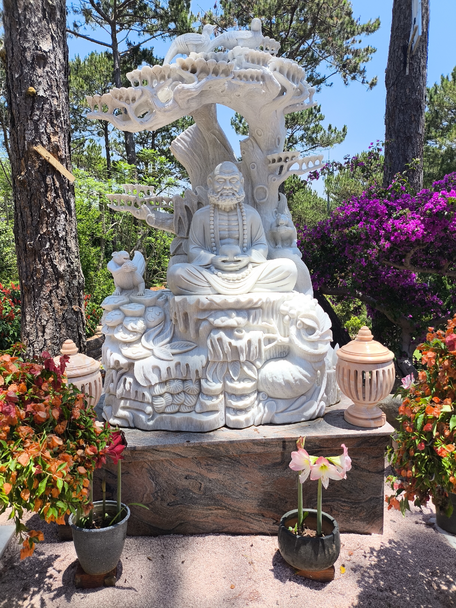 The image depicts a meticulously crafted marble statue, likely located in a serene outdoor setting in Da Lat, Vietnam. The statue features a bearded man seated in a meditative pose, adorned with prayer beads. Surrounding the figure are animal sculptures, including birds and squirrels, all carved from the same white marble. The statue is sheltered by a stylized marble tree canopy. The entire ensemble rests on a dark marble base. Flanking the statue are two ornate, lidded urns made of pale terracotta. Vibrant flower arrangements in hues of orange and red are positioned on either side. In the foreground, potted plants, one with delicate pink and white blooms, add to the peaceful ambiance. Tall trees and purple flowering bushes form the backdrop, bathed in bright sunlight under a clear blue sky.
