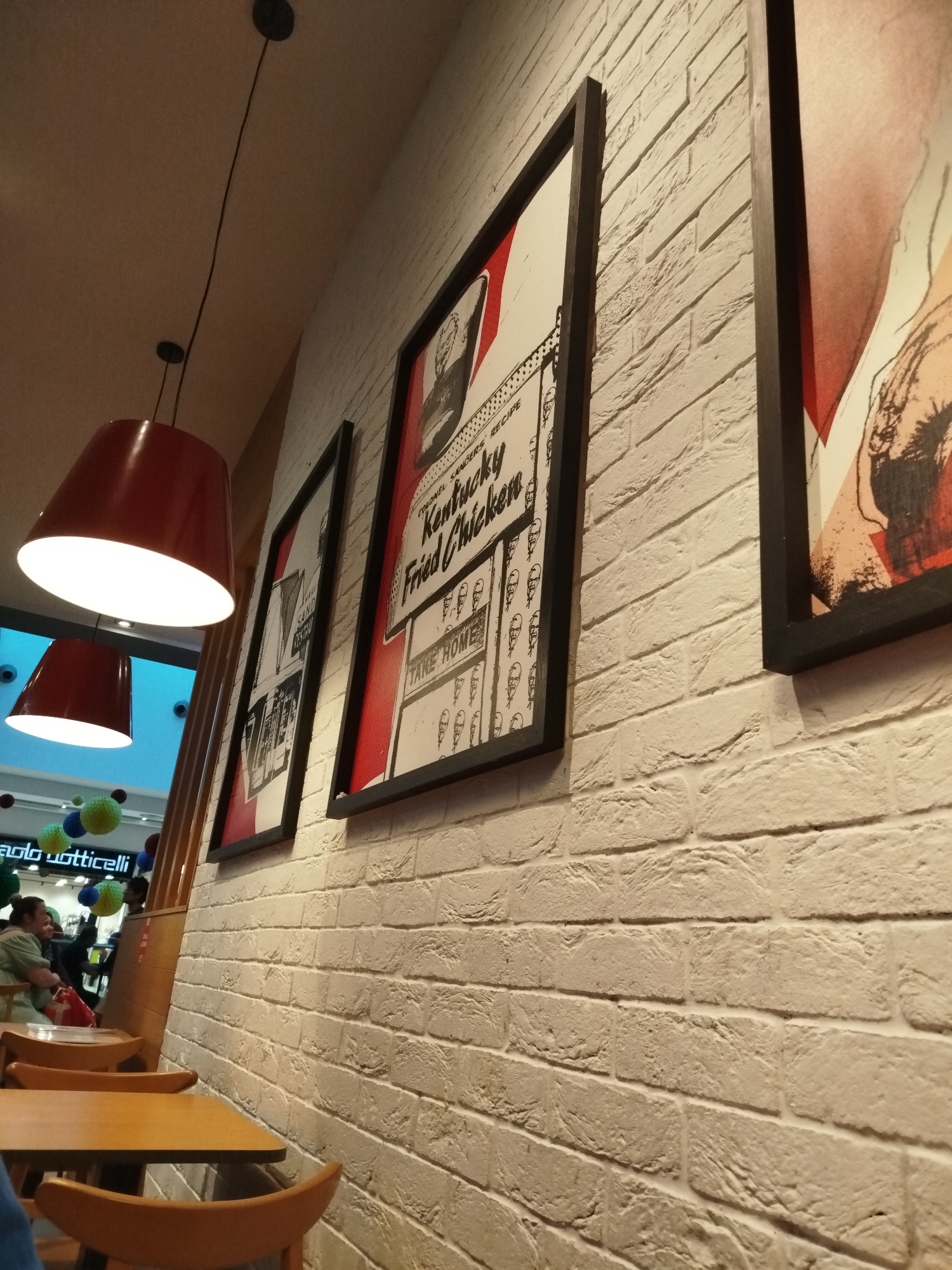 This image depicts the interior of a restaurant or food court, likely a Kentucky Fried Chicken establishment, given the prominent posters. The setting features a dominant white brick wall on the right side, adorned with three framed vintage-style advertisements.
The central poster is clearly visible, featuring "COLONEL SANDERS RECIPE" and "Kentucky Fried Chicken" in a red, white, and black design, along with the text "TAKE HOME" and a graphic of Colonel Sanders. Another similar poster is partially visible to its right, and a third to its left.
In the mid-ground and foreground on the left, several wooden tables and chairs with curved backs are arranged for dining. Two large, red, bell-shaped pendant lights hang from the ceiling, casting bright white light downwards onto the tables. Black electrical cords connect these fixtures to the ceiling.
In the background, beyond the dining area, a brighter, more open space is visible, suggesting a shopping mall or a larger commercial complex. Colorful spherical decorations in green, blue, and yellow hang from the ceiling in this area. A sign with the text "paolo botticelli" is partially visible below these decorations.
Two people are seated at a table in the background; one individual in a light-colored top has a red bag nearby. The scene is well-lit, suggesting either daytime indoors or evening, with artificial lighting dominating. The overall atmosphere appears casual and inviting. This specific location is identified as being in Varna, Bulgaria.