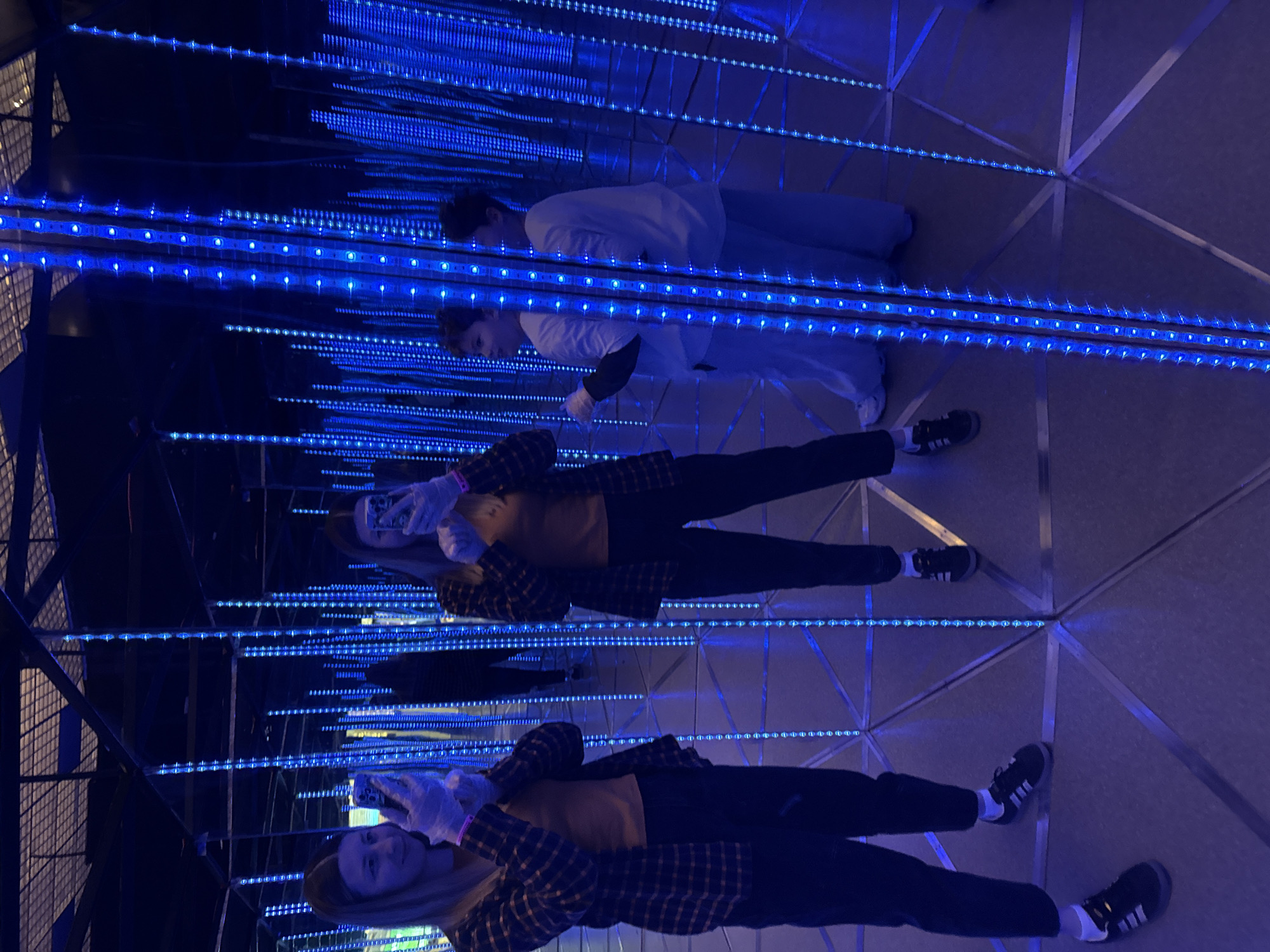 Two people are inside the Museum of Illusions in Moscow, Russia. The room has mirrors and blue lights and is designed to create an illusion of infinite space. One person is wearing black pants and a yellow shirt with a black-and-blue plaid jacket, while the other is wearing a white shirt and light-colored pants. The first person is holding a phone in their hand and is taking a picture or video. The scene is illuminated by the blue LED lights that outline the mirrors. The floor is tiled.