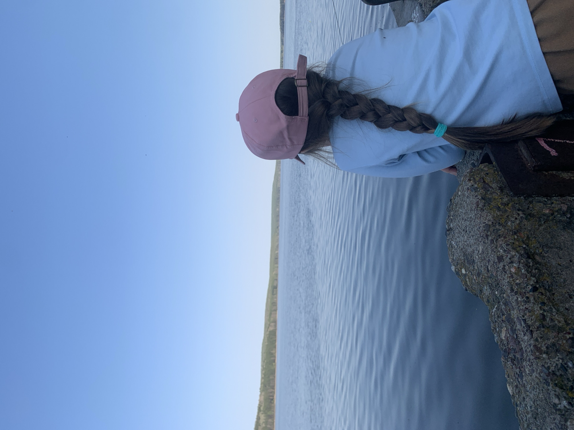 A young person with a pink cap and long braided brown hair, secured with a teal hair tie, is seen from behind, leaning over a concrete structure near a body of water. The person is wearing a light blue long-sleeved shirt and brown pants. The water extends to the horizon, where a thin strip of green vegetation is visible. The sky is a light blue. The photo was likely taken at the city of Chaikovsky, Russia, based on the request. The person is likely observing something in or near the water.