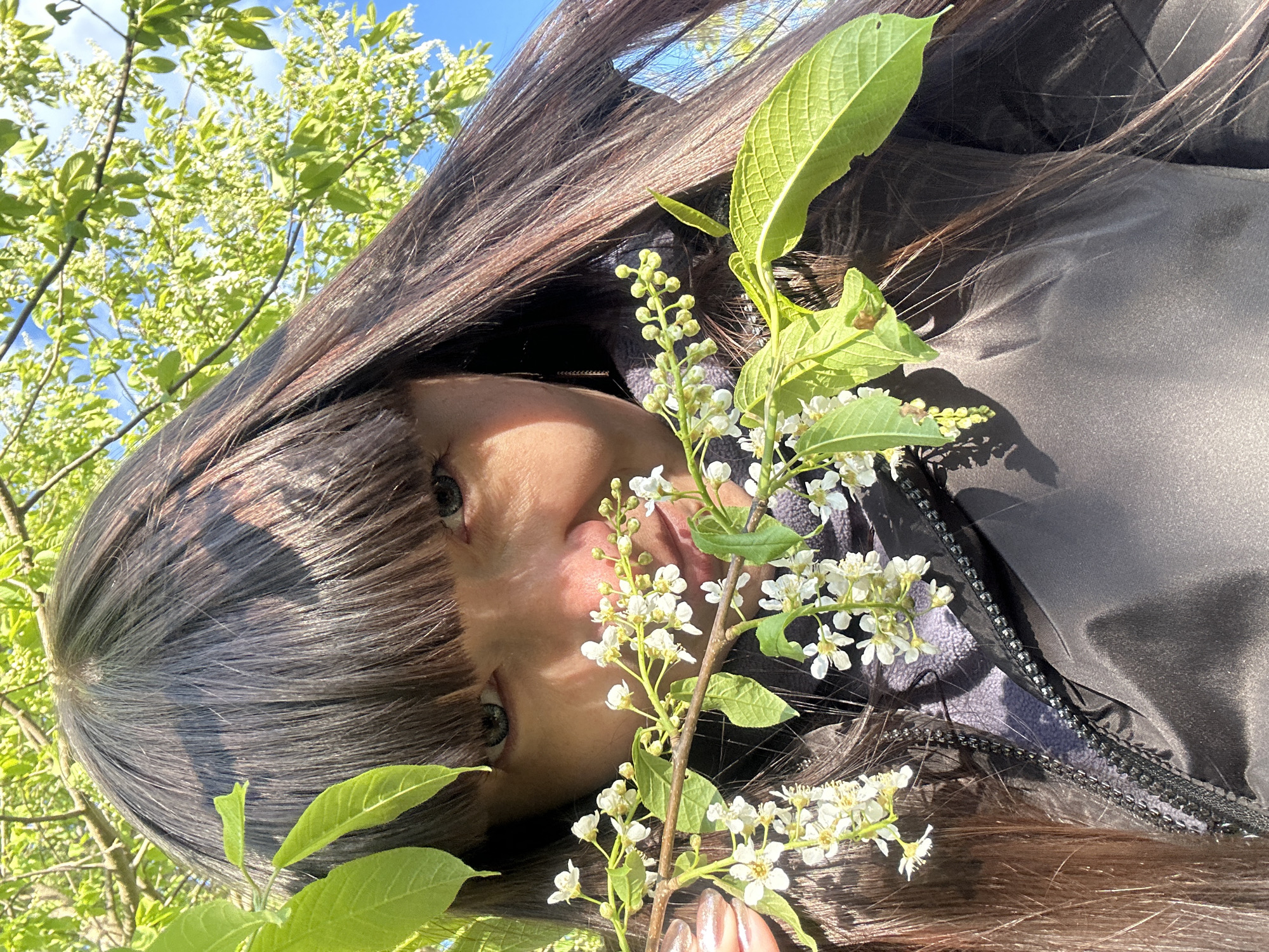 A woman with dark brown hair and bangs looks up at the camera, her face framed by a branch of white flowers and green leaves. She is wearing a dark-colored jacket with a visible zipper. The background consists of green trees and a blue sky. The image appears to be taken outdoors in a natural setting with lush greenery. It's impossible to confirm the location as Среднее Кечево, Russia based on the image alone.