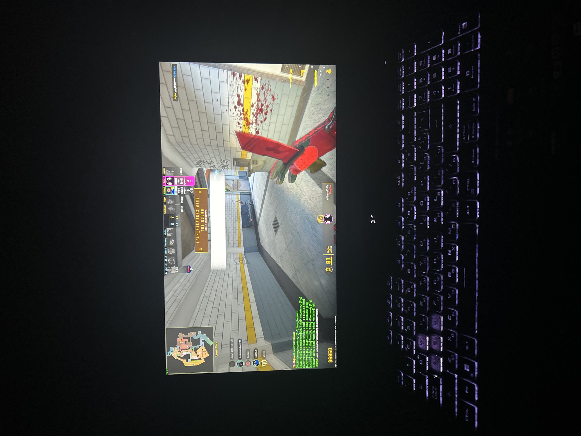 The image shows a monitor displaying a first-person perspective of the video game Counter-Strike: Global Offensive (CS:GO). The game is set on the map "Lower Park," with the player's in-game avatar wielding an Ursus Knife with the "Slaughter" skin. Blood splatters are visible on the wall. In-game UI elements such as team scores, player statistics, and money are displayed. Text chat is present. A backlit keyboard is visible in the lower right of the frame. The "Lower Park" map is a custom-made map for CS:GO, and is NOT connected to any specific real-world location in Varna, Bulgaria or anywhere else.