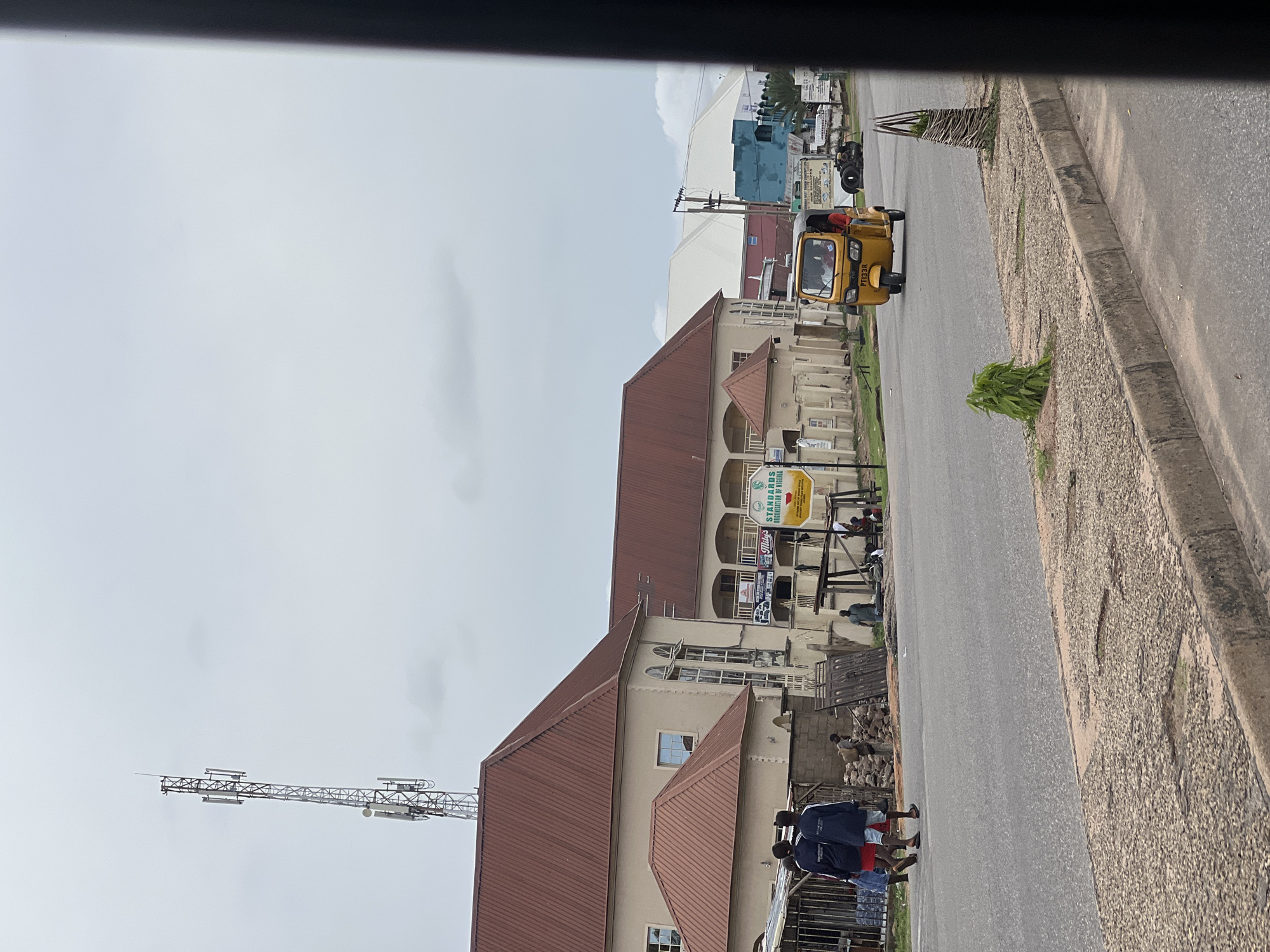 The image depicts a street scene in Jalingo, Nigeria, under an overcast sky. On the left, a two-story building with a reddish-brown corrugated metal roof and cream-colored walls is visible. A tall communication tower stands prominently behind this building. In front of the building, two individuals, likely young men, are walking on the side of the road; one is wearing a red t-shirt and shorts, and the other is in a dark blue jacket and shorts. A busy street with gray asphalt stretches across the foreground and middle ground. A yellow auto-rickshaw, also known as a "Keke" in Nigeria, is traveling down the street on the right side of the frame, driven by a person in a red shirt. Several other buildings and structures are visible in the background, including a white building with a light-colored roof and a blue building. The road has a worn asphalt surface with a concrete curb and gravelly shoulders. Signs are visible on the buildings, one of which reads "STANDARDS ORGANIZATION OF NIGERIA." Construction materials, including wooden planks and piles of stones, are present on the side of the road near the buildings.