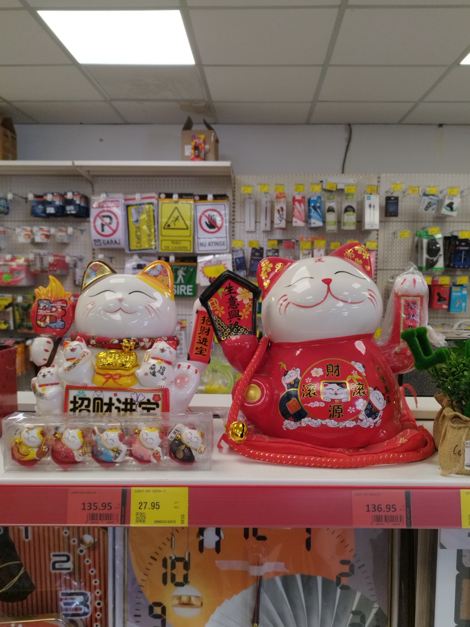 The image shows a retail store shelf in Bucharest, Romania. On the shelf are two large ceramic lucky cats, also known as Maneki-neko, a popular Japanese figurine. The cat on the left is white with gold accents, and the cat on the right is red with gold and black accents. Both cats are depicted with one paw raised in a beckoning gesture, a characteristic of these figurines. In front of the larger cats, on the same shelf, is a clear plastic package containing a set of five smaller, similarly designed lucky cat figurines. Below the shelf, price tags are visible, indicating prices of 135.95 lei and 27.95 lei, and 136.95 lei, for the lucky cat items. The background of the image shows shelves stocked with various retail items, including packaged electronics and signage. The environment appears to be a well-lit store with fluorescent ceiling lights.