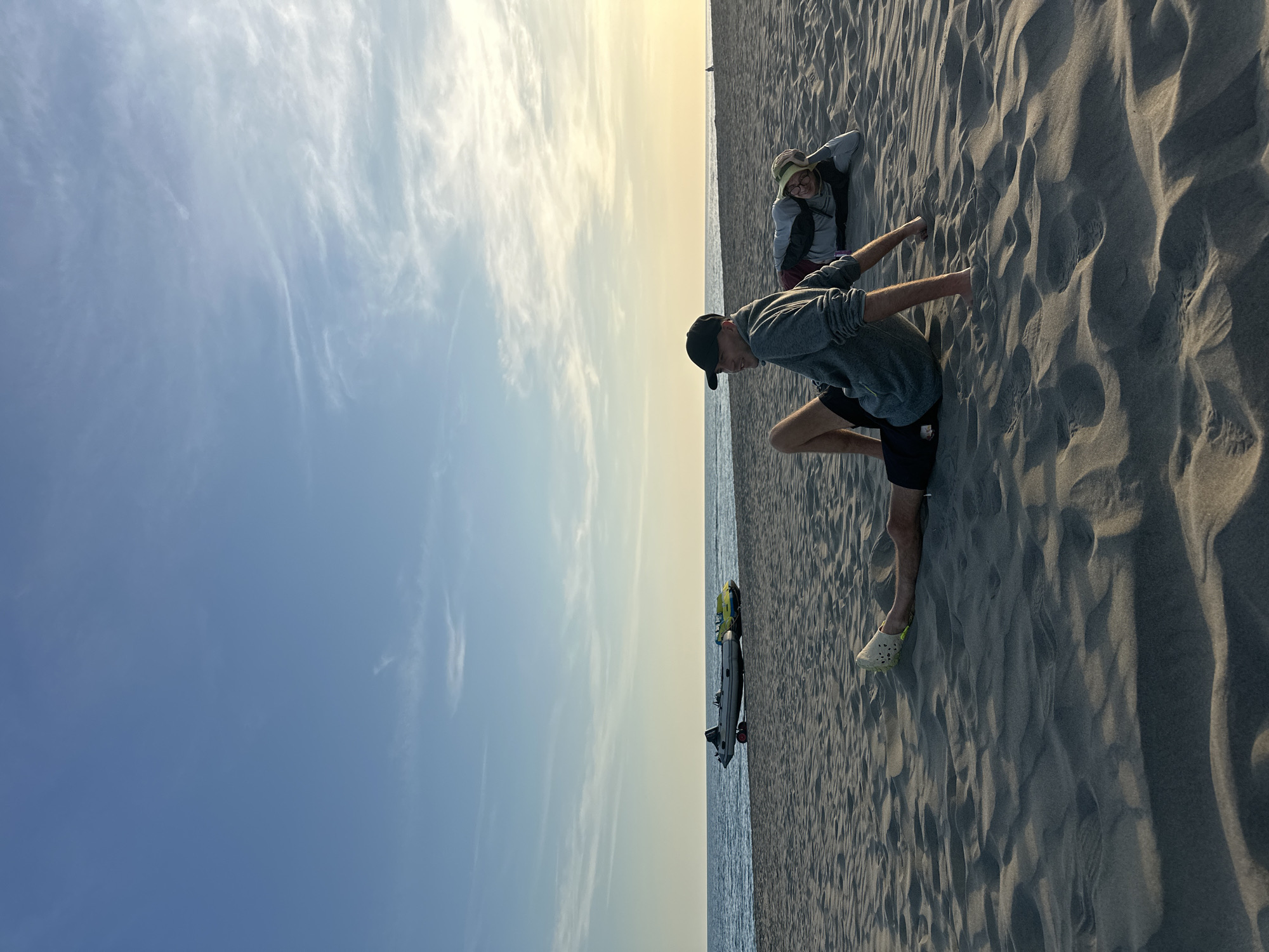 The scene is a beach landscape in Donji Štoj, Montenegro. The photograph shows two individuals on a sandy beach with the sea and a light blue sky visible. The person in the foreground is a young male wearing a black cap, grey hoodie, black shorts, and light-colored footwear. He is crouching with his hands touching the sand. Further behind him, a second person is lying down on the beach, with their head resting on their arms. Near the sea, there is a personal watercraft parked on the sand.