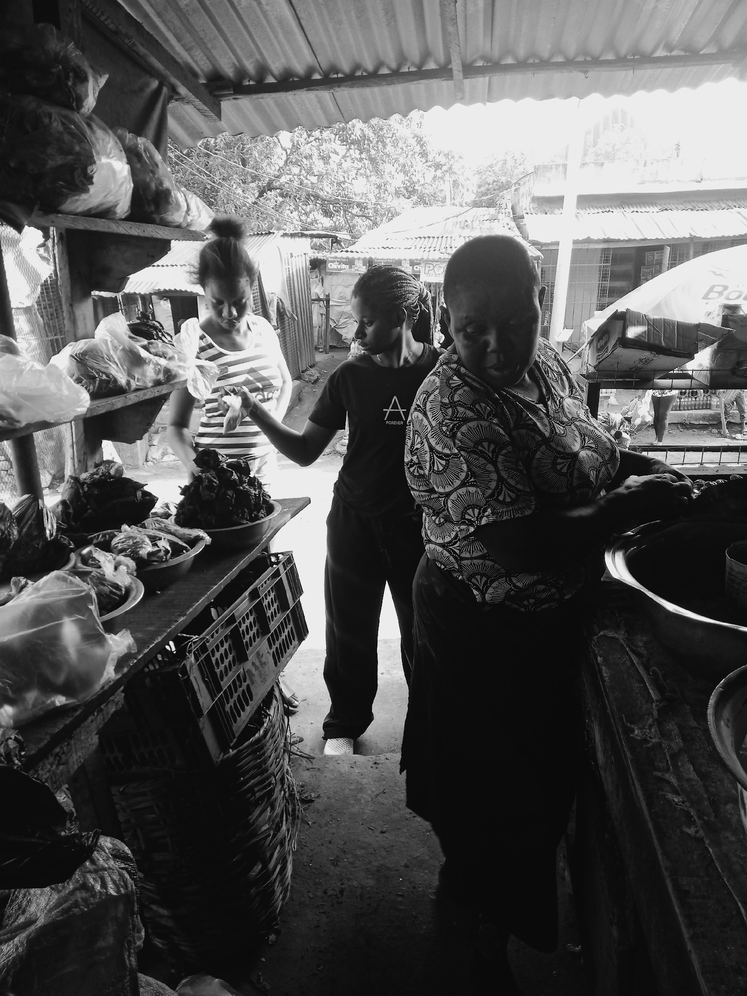 Here is a description of the image:
The image is a black and white photograph taken indoors, appearing to depict a market scene in an urban setting. Three women are the main subjects.
The woman on the right stands closest to the camera, partially obscured in shadow from the waist down. She is wearing a patterned, short-sleeved shirt. Her attention is focused downward, seemingly working with something in a large metal bowl or pot in front of her.
To the left of her stands another woman, wearing a dark t-shirt with a visible white logo and text. She is extending her arm towards a woman who is looking down at food and is wearing a striped short-sleeved shirt and has her hair pulled into a bun.
The setting is a simple market stall or shop. In the background, glimpses of other structures, possible houses, and trees can be seen. Overhead, a corrugated metal roof provides shelter. A table laden with bowls of food and packaged goods is to the left. There is some storage under the table, including a crate and a wicker basket filled with bags.