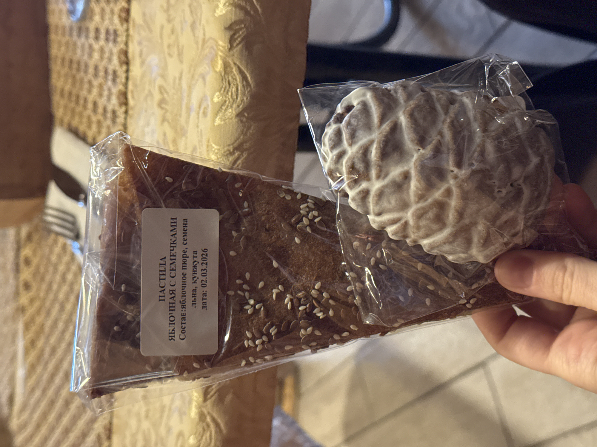 A close-up shot shows two types of individually packaged food items held in someone's hand. The one on the left, a triangle-shaped piece of apple pastila with seeds, is partially covered by a label with Russian text that reads "Apple pastila with seeds, ingredients: apple puree, flax seeds, sesame seeds, date: 02.03.2026." The other item is a round, cookie-like treat with a white, textured surface. Both are sealed in clear plastic packaging. The background is slightly blurred, showing the corner of a dining table with silverware and a patterned chair. There is no mention of Ново-Никольское, Russia in the image.